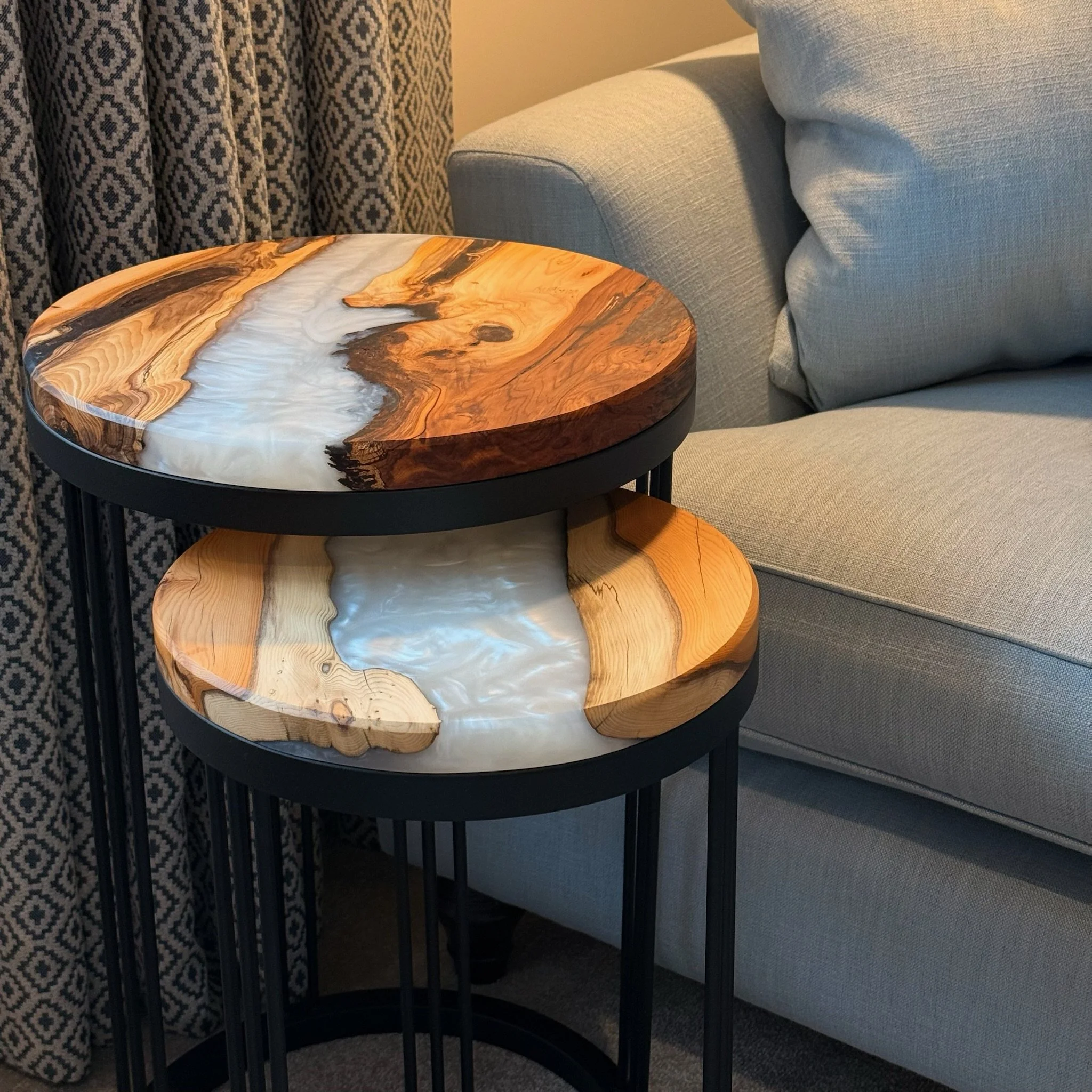 A set of two overlapping round side tables with wood and resin tops next to a gray sofa and patterned curtains.