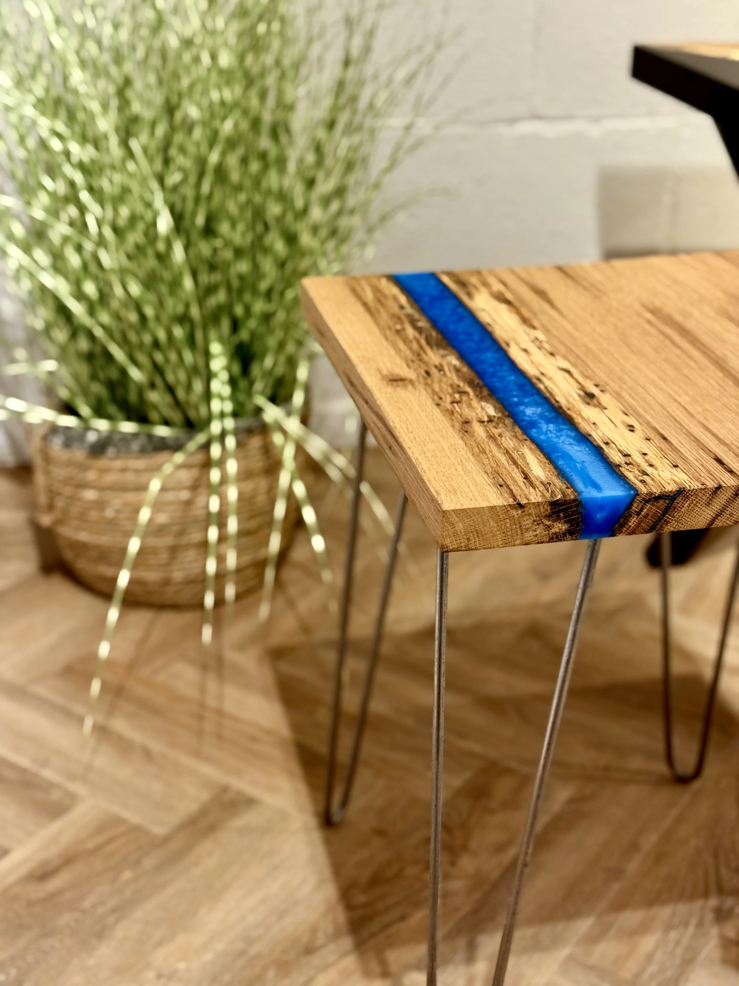 Close-up of a wooden side table with a blue resin inlay on top, resting on metal hairpin legs, with a large green plant in a woven basket in the background.