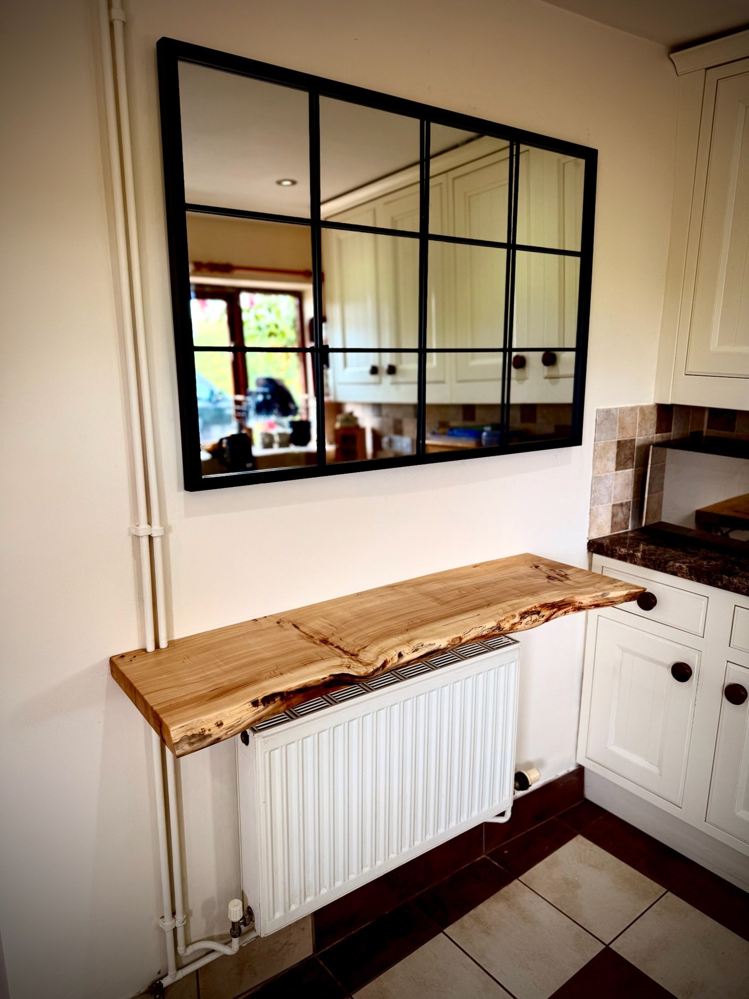 A wooden side table with a natural edge sits above a white radiator in a kitchen, with a mirror reflecting kitchen cabinets and a window with outside scenery.