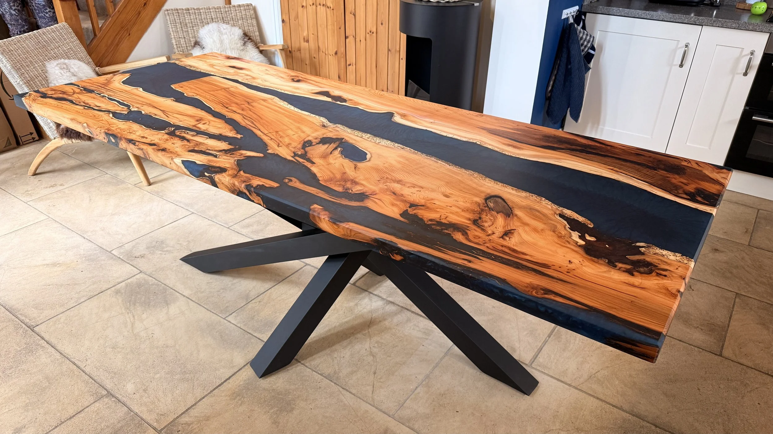 A wooden table with a black adhesive resin inlay on a black X-shaped metal base, situated on a tile floor in a room with kitchen cabinetry and wood-paneled walls.