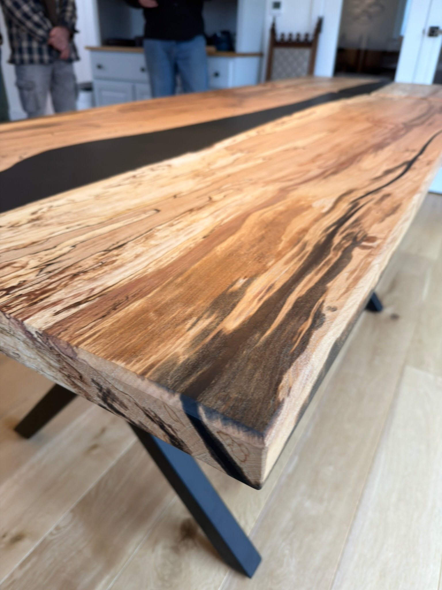 Close-up of a wooden table with a black resin inlay running along the center. The table has a natural, rustic finish with visible wood grain and a live edge.