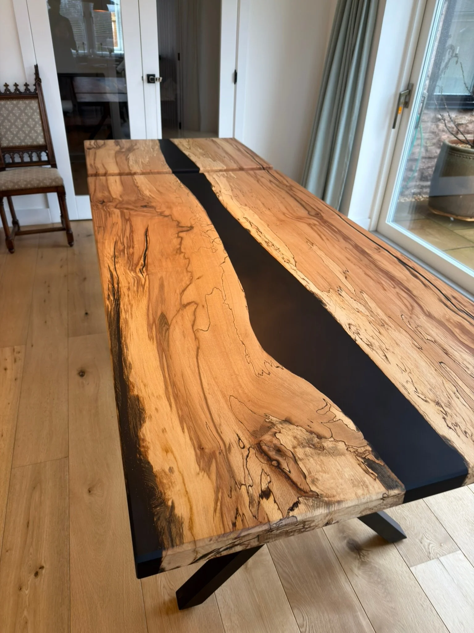 A wooden table with a natural edge and black resin inlay running through the center, situated in a room with wooden flooring and sliding glass doors.