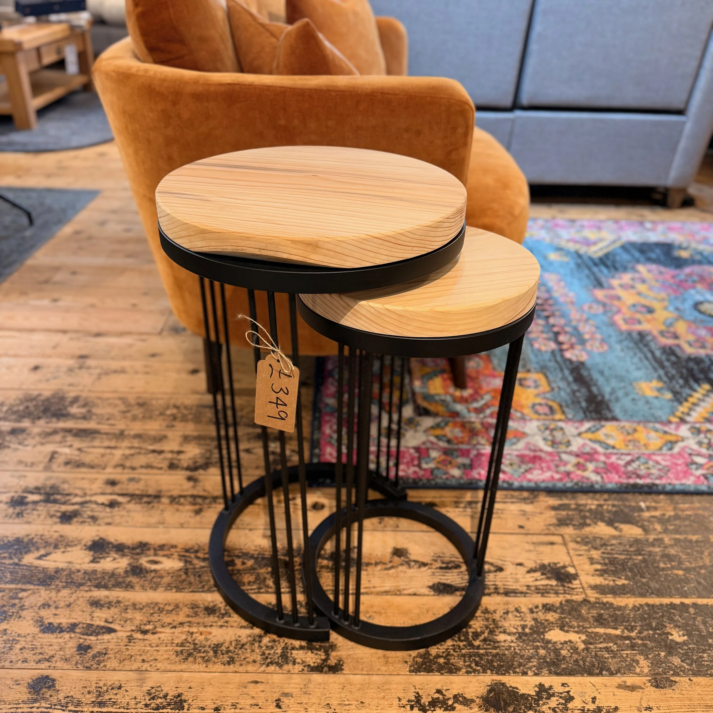 Two nesting side tables with round wooden tops and black metal bases, placed on wooden flooring near an orange upholstered armchair.