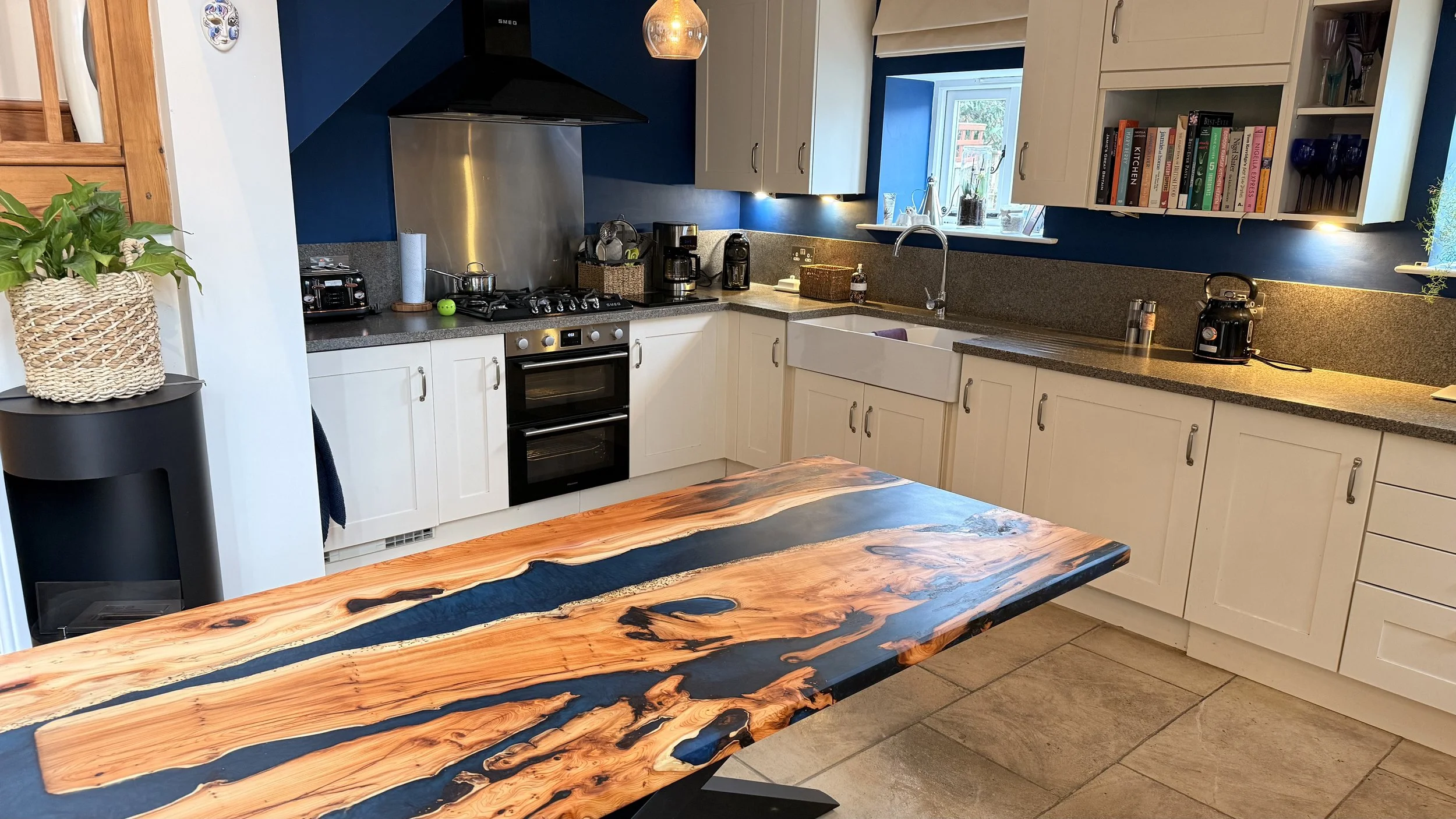 A kitchen with white cabinets, a black stove with double ovens, a window above the sink, and various appliances on the countertops. There is a wooden table with a glossy finish in the foreground.