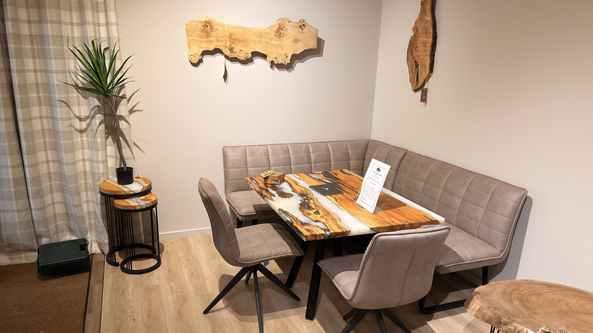 A dining area with a rectangular wooden table with orange, black, and white marbled patterns, surrounded by a beige upholstered corner bench and four matching beige chairs. On the wall above the table are two pieces of natural wood art. To the left, 