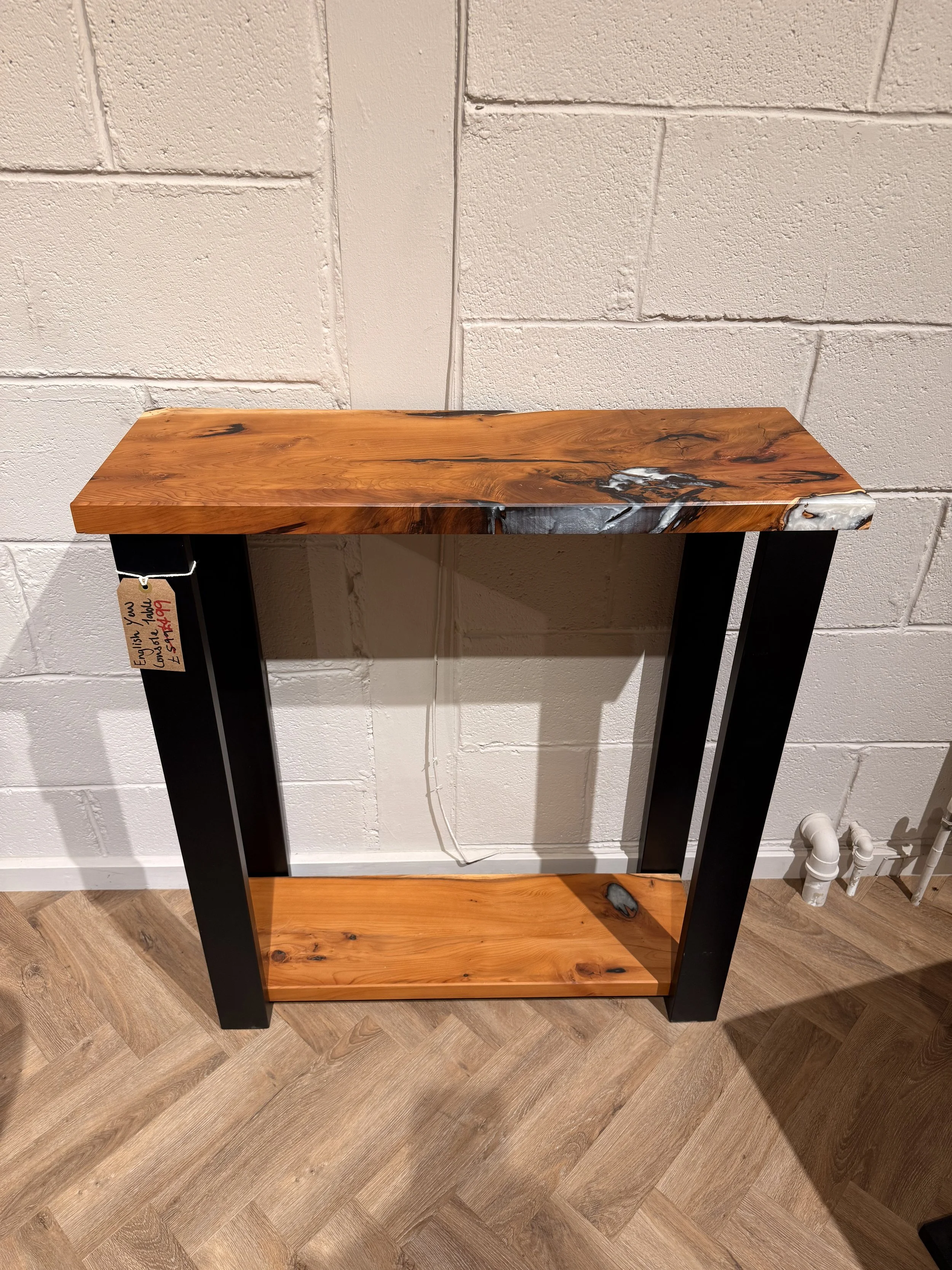 A wooden and black metal console table placed against a white brick wall. The table has a wooden top and base with black metal supports on the sides.