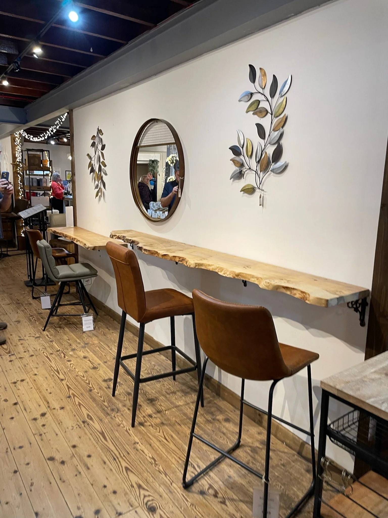 A display of a wooden floating bar-style table with three chairs against a white wall in a retail store, with decorative wall metal leaves and a round mirror, hardwood floor, and ceiling track lighting.