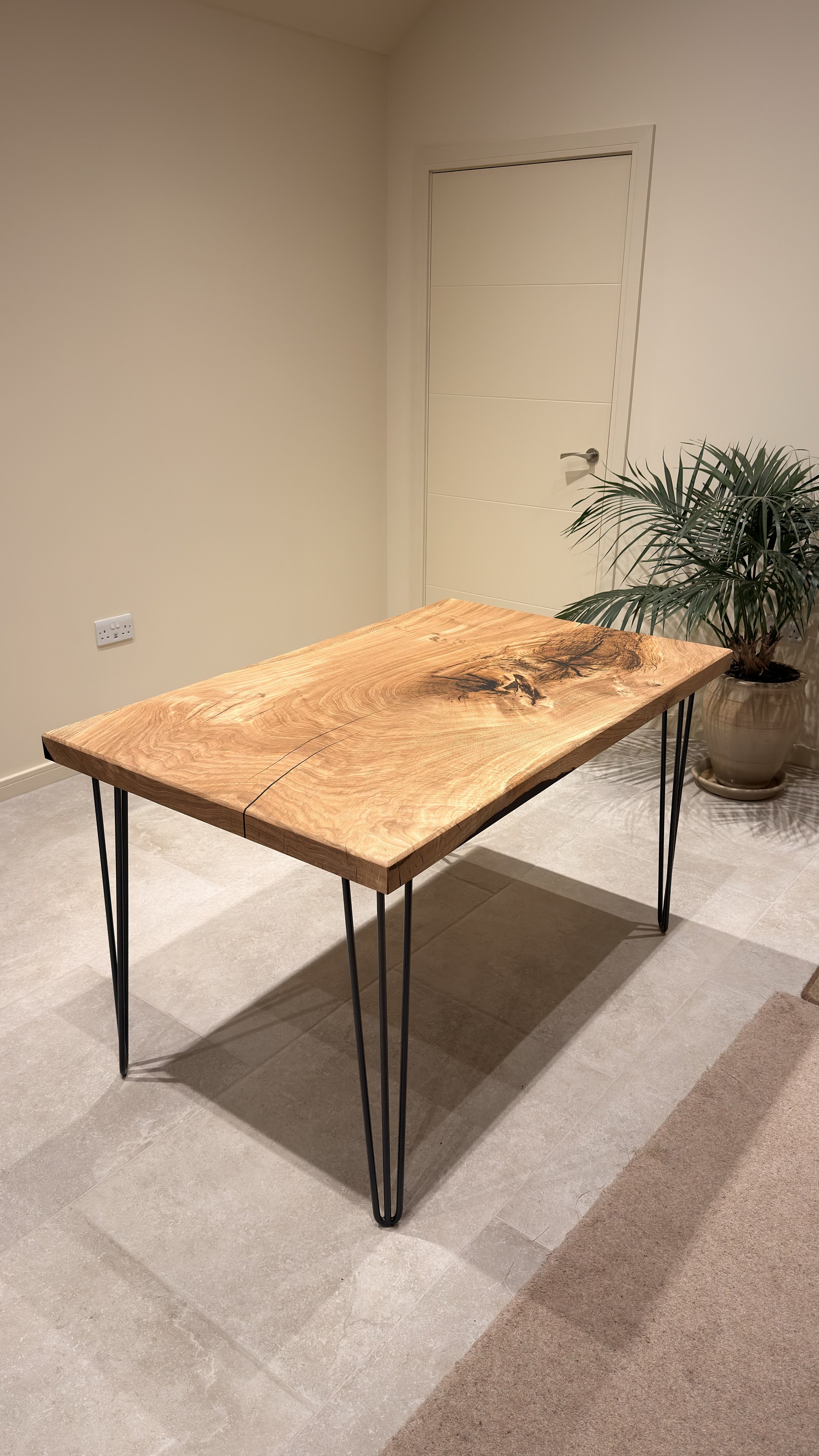 A wooden table with metal hairpin legs in a room with beige walls, a door, a power outlet, a potted plant, and beige tiled flooring.