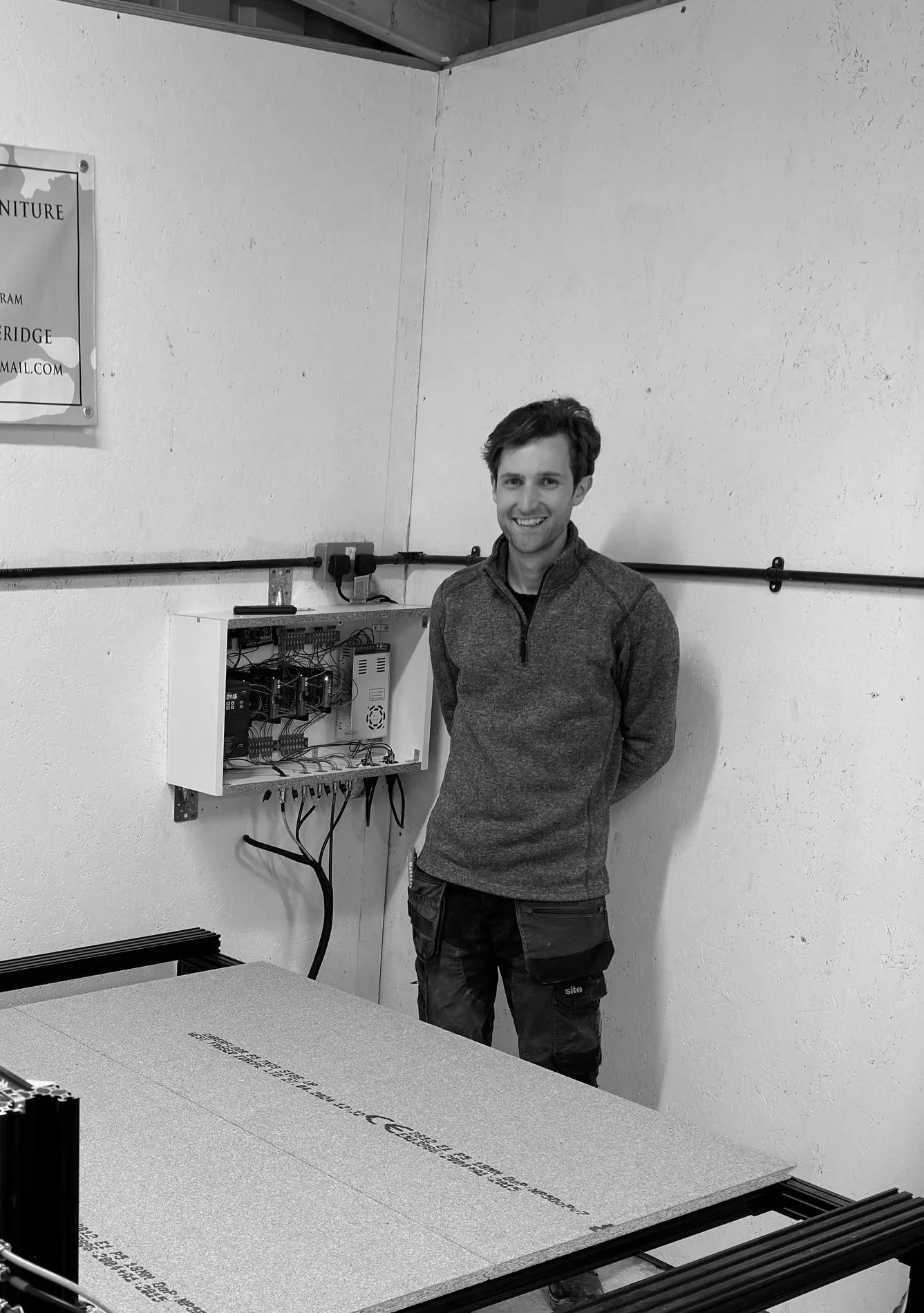 A smiling man in a work jumpsuit standing indoors next to a woodworking project with clamps, with a background of trees and a vehicle outside.