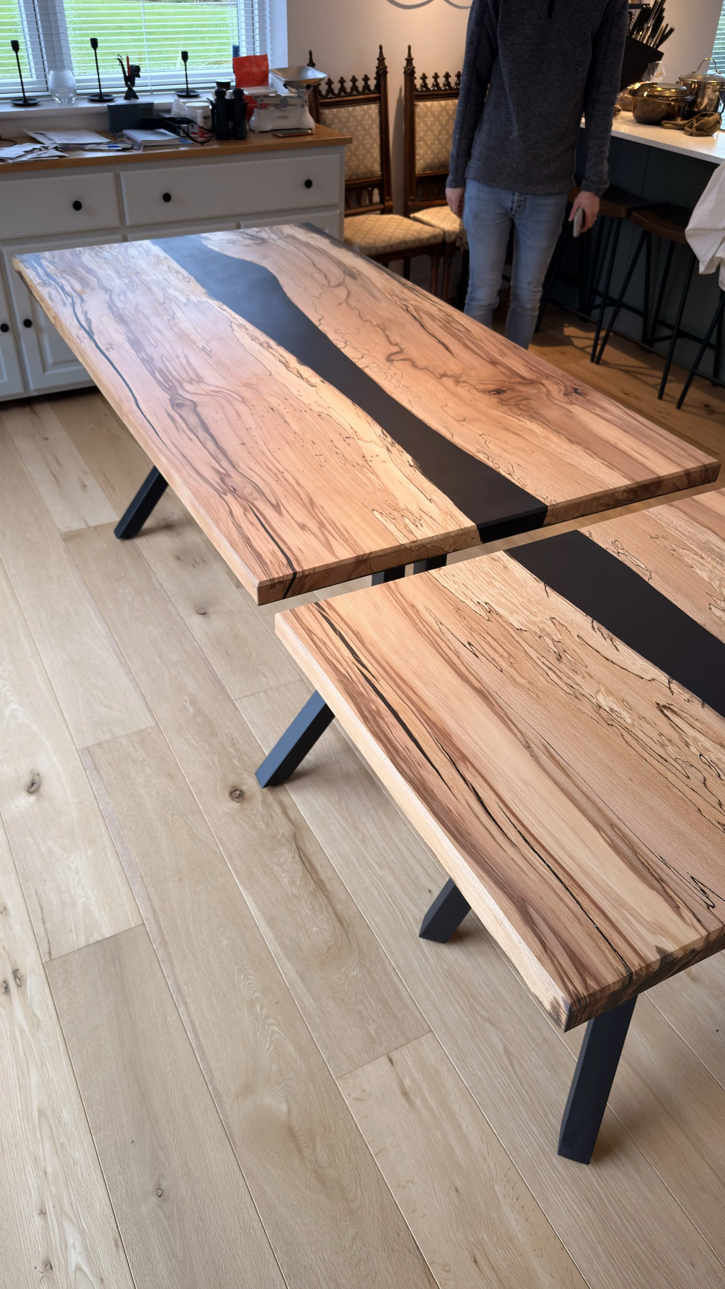 Wooden table with a black resin strip in the center, supported by black angled legs, placed on a light wooden floor in a room with a window and various objects on a sideboard.
