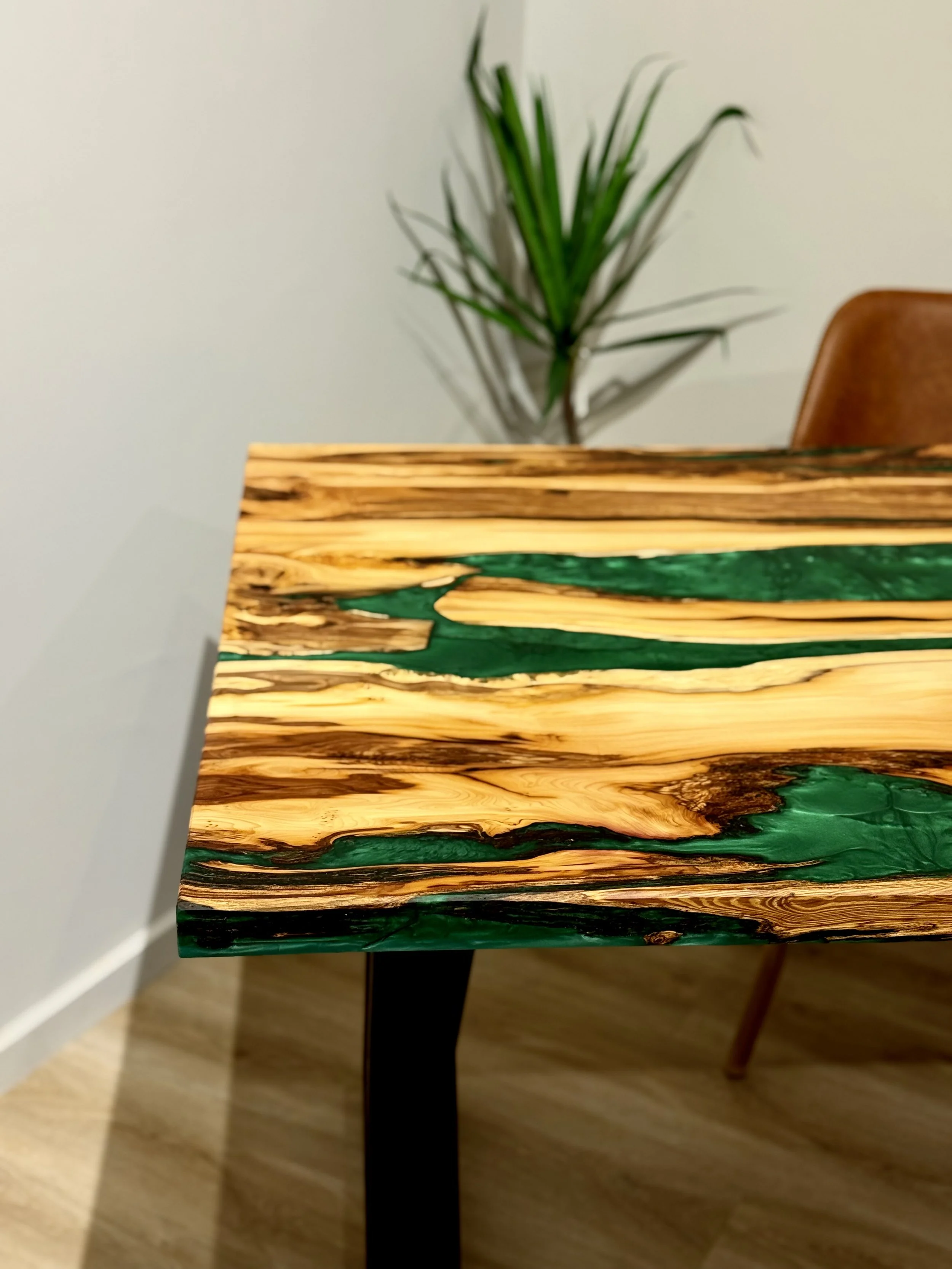 Close-up of a wooden table with a glossy finish and green resin accents, in a room with a plant and a chair in the background.