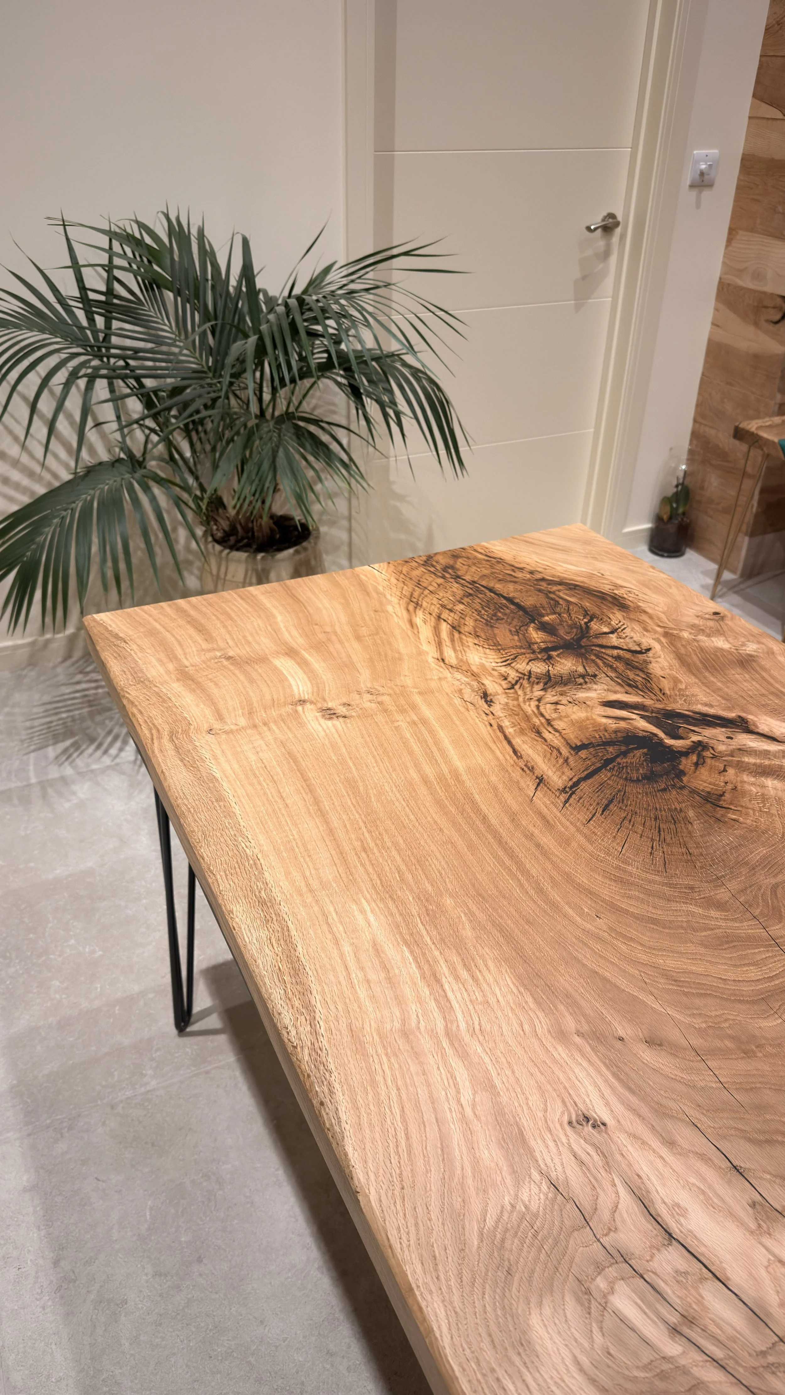 Close-up of a wooden table with visible wood grain and knots, with a green potted plant in the background near a white door and wooden wall.