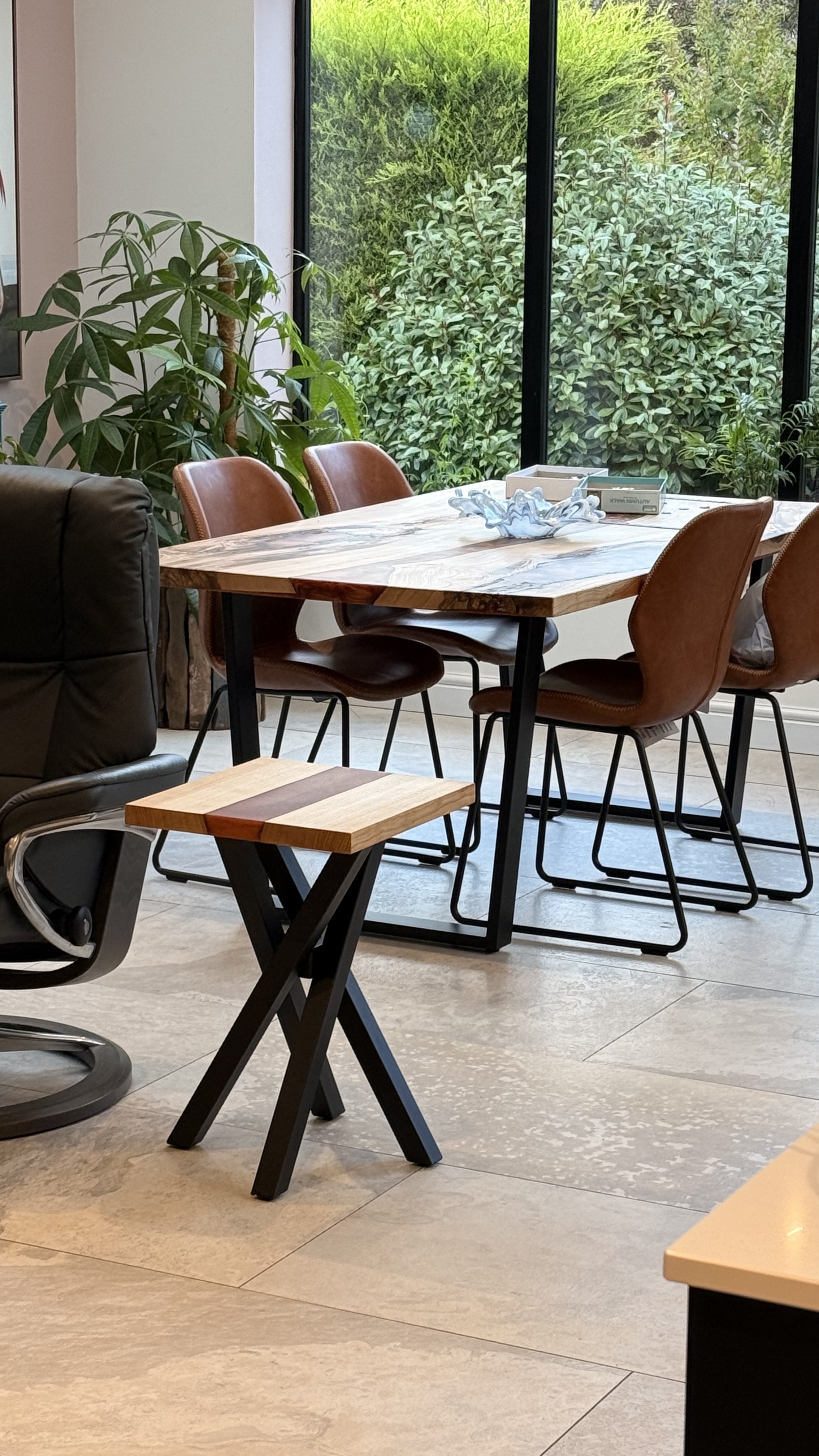 A dining area with a wooden table and four brown chairs near large windows with green trees outside. There's a small wooden side table in the foreground and an indoor plant beside the window.