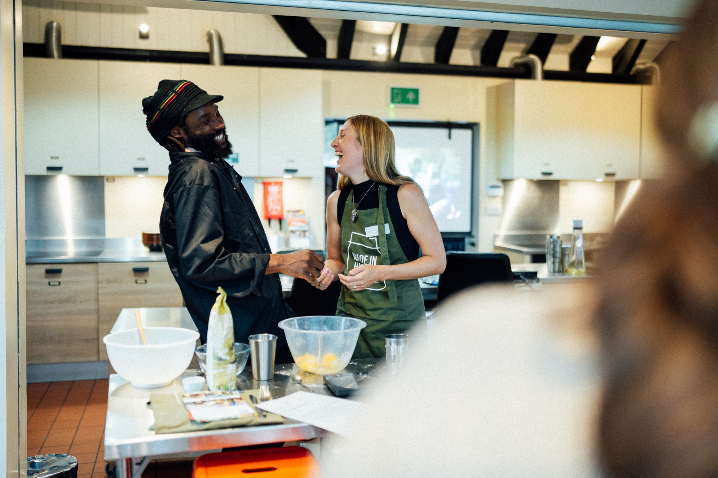 Two people are laughing and talking in a kitchen. The man has dreadlocks and a black beanie, and the woman has blond hair and is wearing a green apron. There are bowls and utensils on the kitchen counter.