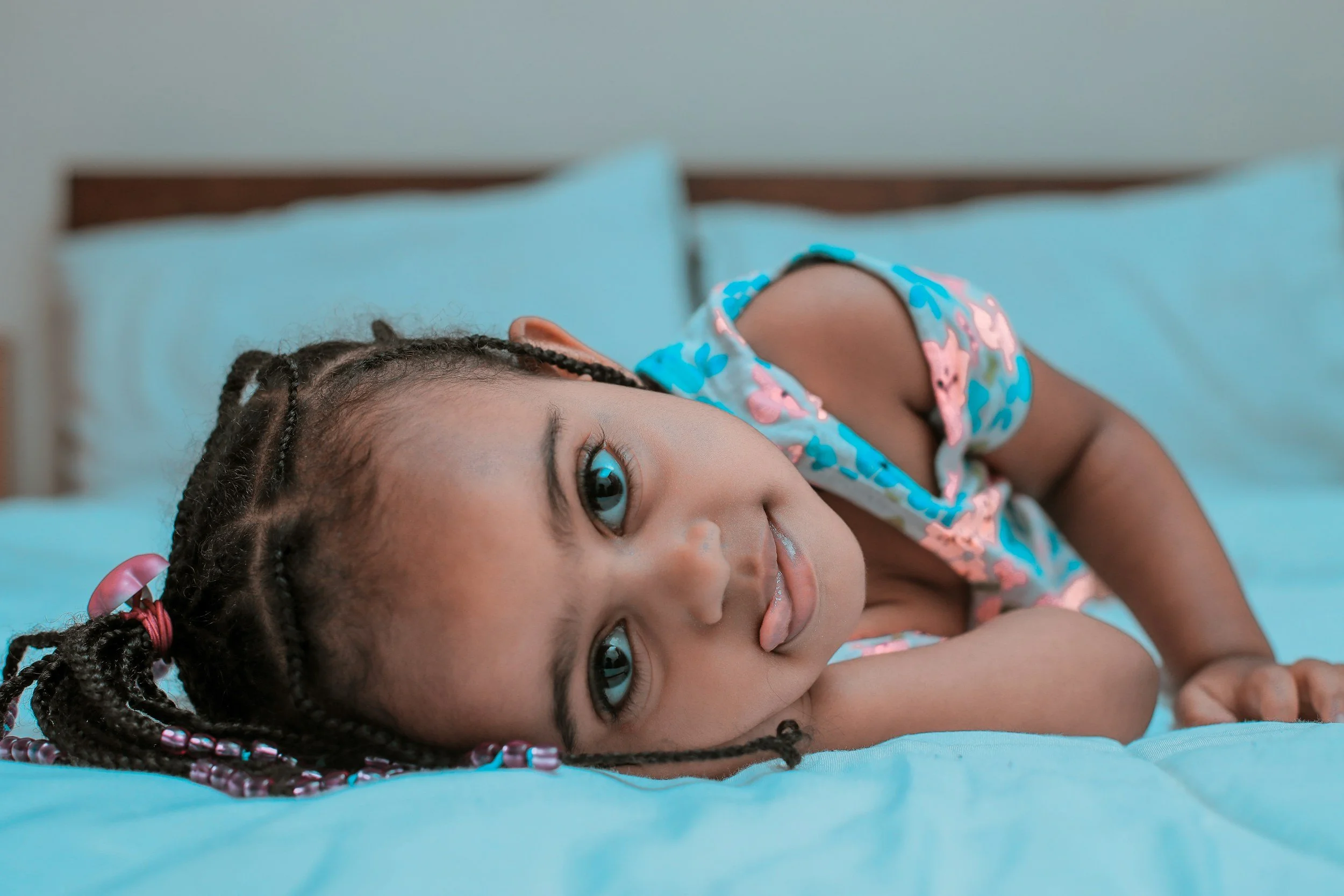 Close-up of a young girl lying on her side on a bed with turquoise sheets and pillows, wearing a colorful pajama top, with braided hair adorned with beads and pink hair ties, looking at the camera.