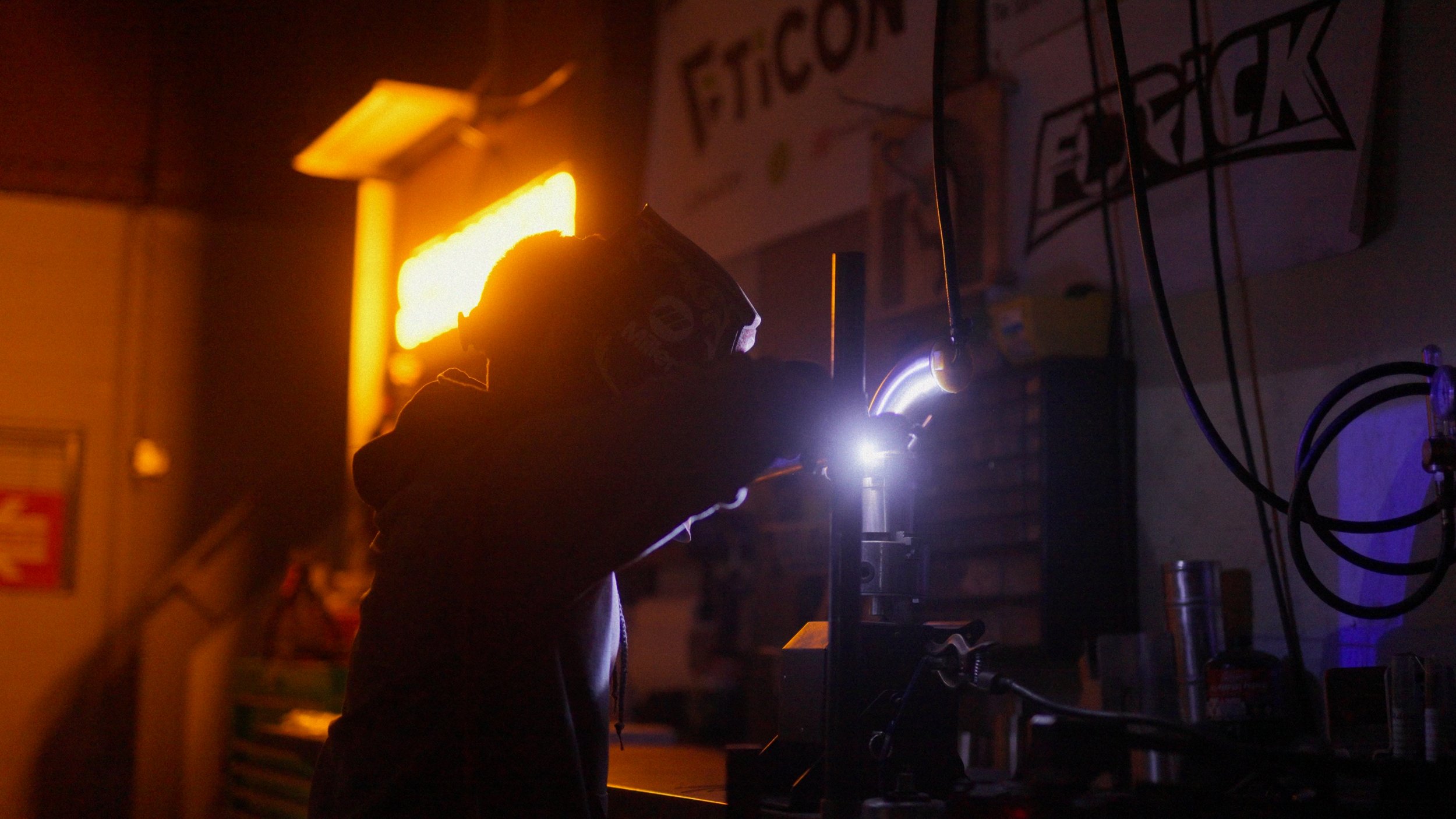 A person welding in a workshop, wearing safety goggles, with a bright arc of light from the welding process illuminating their silhouette, surrounded by various tools and equipment.