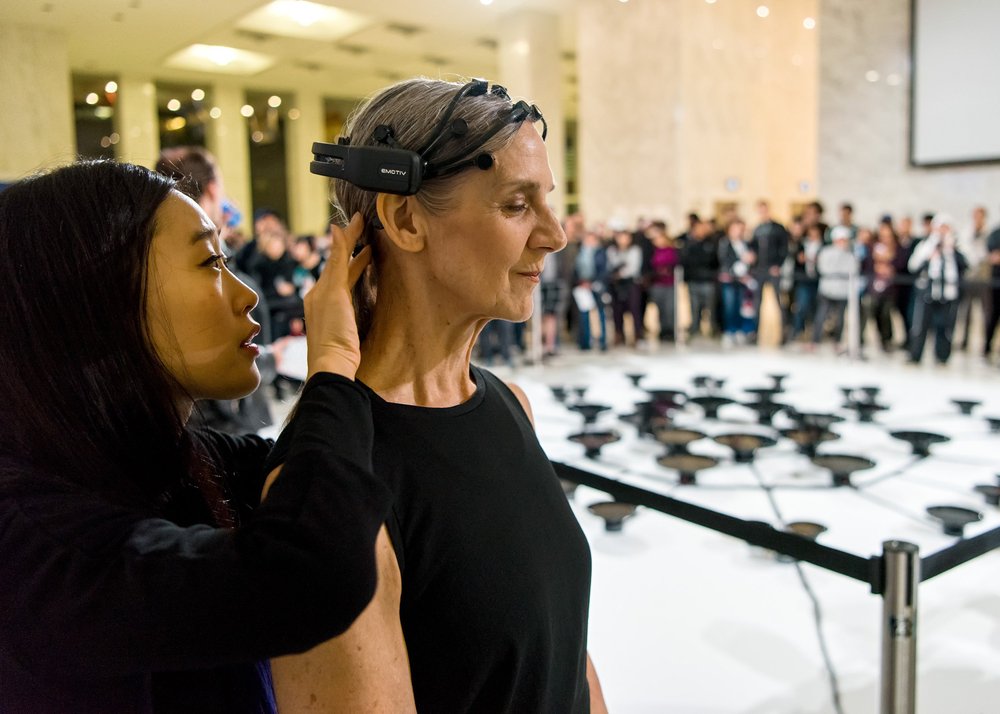  Peggy Baker and Lisa Park, standing just outside of their performance space, a large crowd and the shallow pans holding water in front of them. Lisa is fitting the headpiece equipped with sensors to Peggy’s head. 