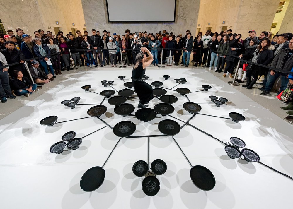  Peggy Baker, resting her arms on her head, standing in the centre of three concentric rings of shallow pans. The ring closest to Peggy has eight pans forming a tight circle, the middle ring is farther from centre with eight pans that are spread apar