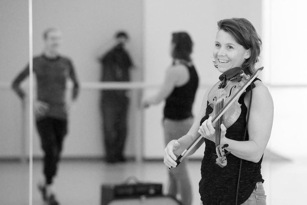  Sarah Neufeld holding her violin against her shoulder and chin, pausing between practice. She is smiling at someone off-camera. In the background, reflected in a mirror, are Sarah, Peggy Baker and the photographer taking the shot. 