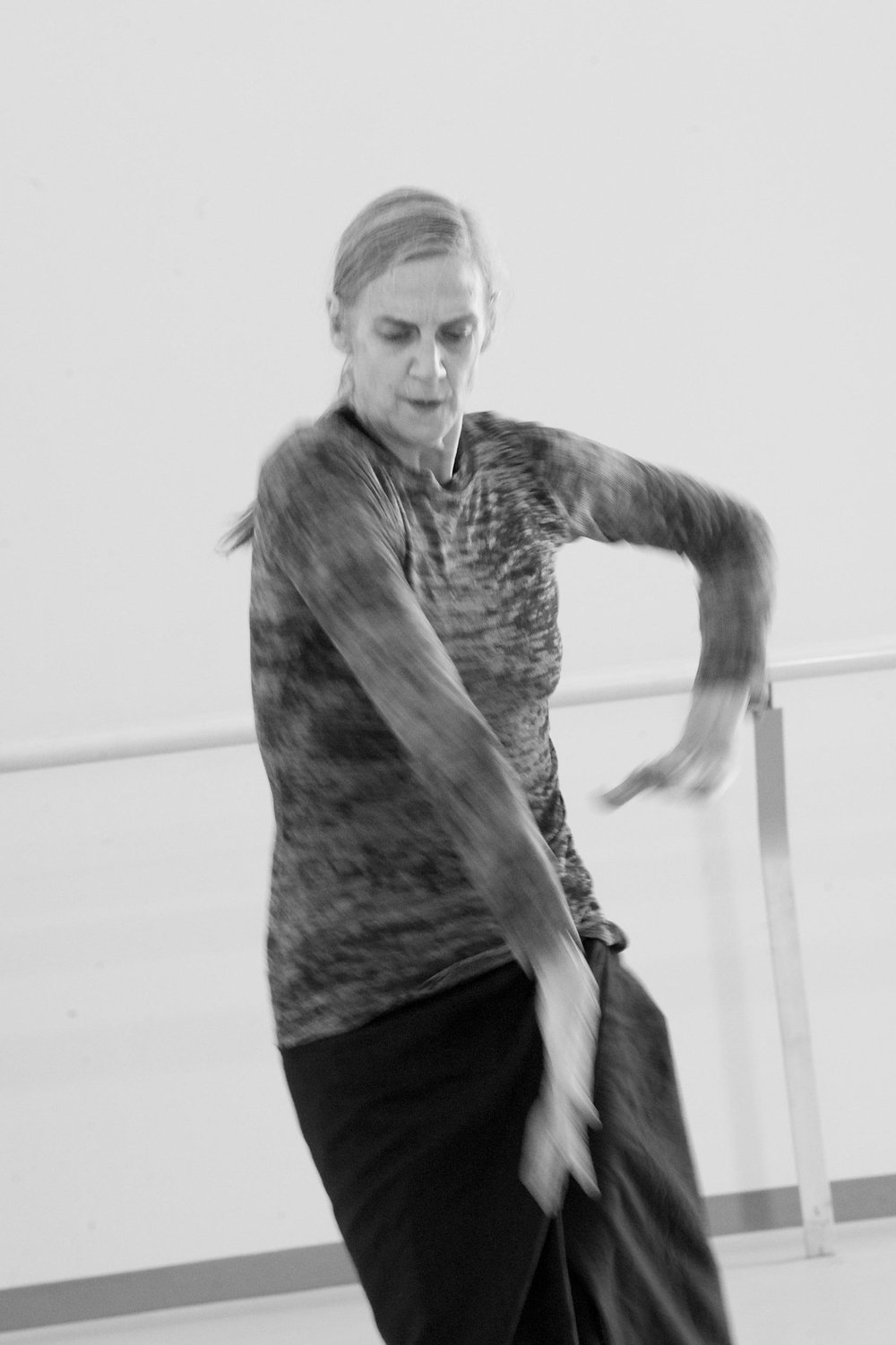  Peggy Baker in a close-up, caught mid-movement. Her right arm is moving straight down to point at the ground in front of her as her left moves to go toward it. Her gaze is down and her hips lead off balance to the left. 
