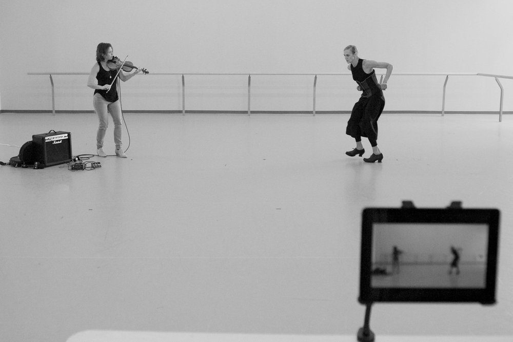  Sarah and Peggy rehearse together. On the left, Sarah is standing and playing her violin, while on the right, Peggy is bent at the knees, her arms bent back with the elbows lifted. In the lower right corner, a camera monitor is visible, recording th