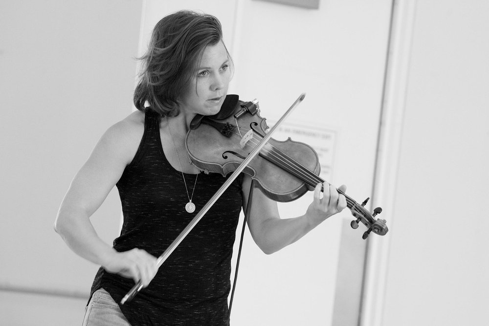  Sarah Neufeld laying her violin during rehearsal. A close-up of her upper body. She is looking off-camera and her bow is mid-movement.  