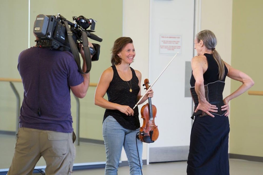  On the left, Sarah and a camera operator. Sarah is speaking to Peggy, who is on the right, in front of Sarah, standing with her hands resting on her lower back. Their conversation is being filmed. Sarah is holding her violin and bow. Both women are 