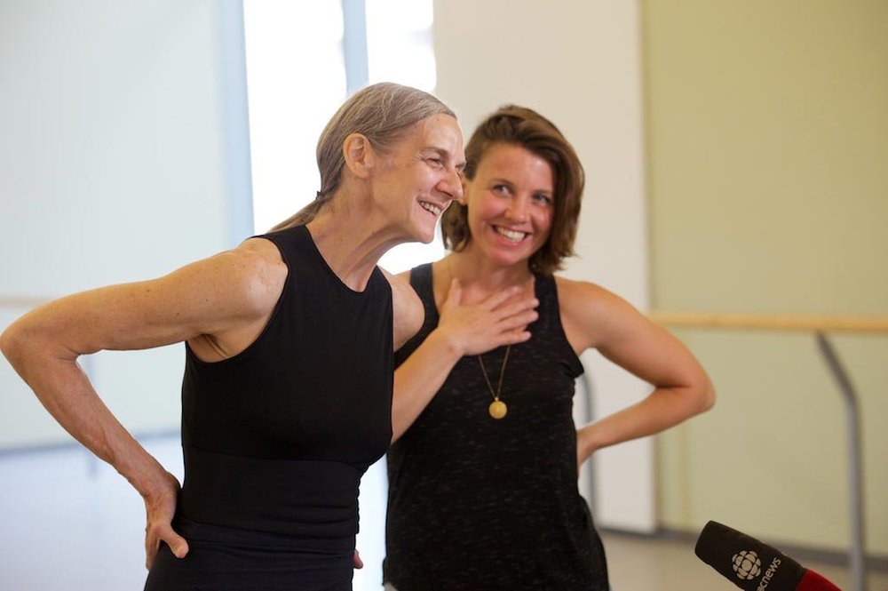  On the left, Peggy Baker speaking to a CBC interviewer off-camera. Her hands are resting on her lower back. To the right and slightly behind her, Sarah Neufeld stands looking at Peggy and smiling with her right hand resting on her collarbone.   
