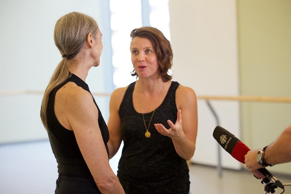  Sarah and Peggy during a CBC interview. They are standing in their rehearsal studio, speaking to each other. Peggy’s back is facing the camera while Sarah is seen gesturing toward her. A CBC microphone is visible in the lower right corner.  