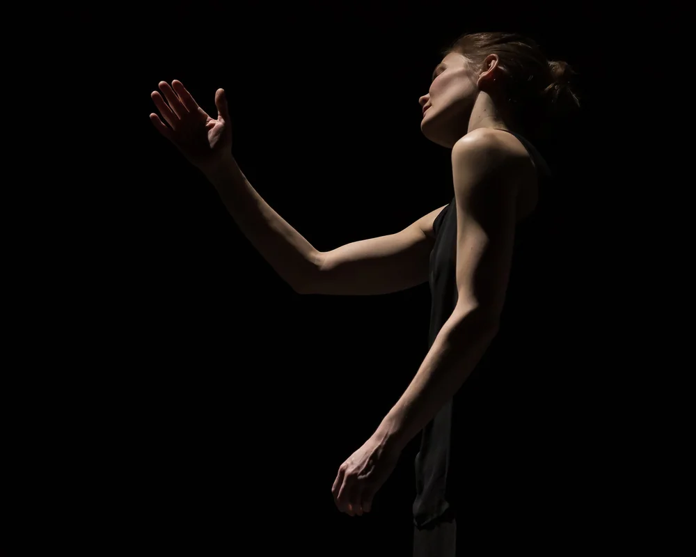  Kate Holden posed with her left side facing the camera. Her right arm is bent and held in front of her face as she gazes at it.&nbsp;She is on a dark stage, and only the edges of her body are lit. 