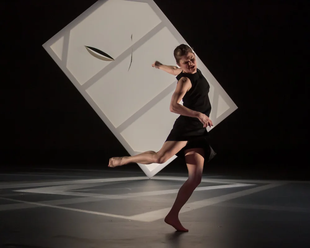  Kate Holden wearing a sleeveless black shift dress is mid-spin. She is on the ball of her left foot with her right foot raised behind her and her left arm is thrown open and back. Se looks down to the right with an intense expression on her face. 