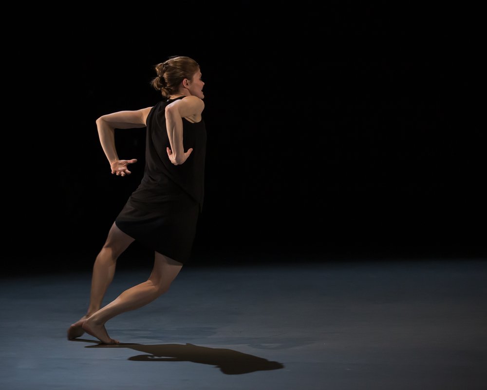  Kate Holden, mid-movement and leanly strongly forward. She is on the balls of her feet, poised as if she is about to run with her arms behind her back, palms facing upwards. 