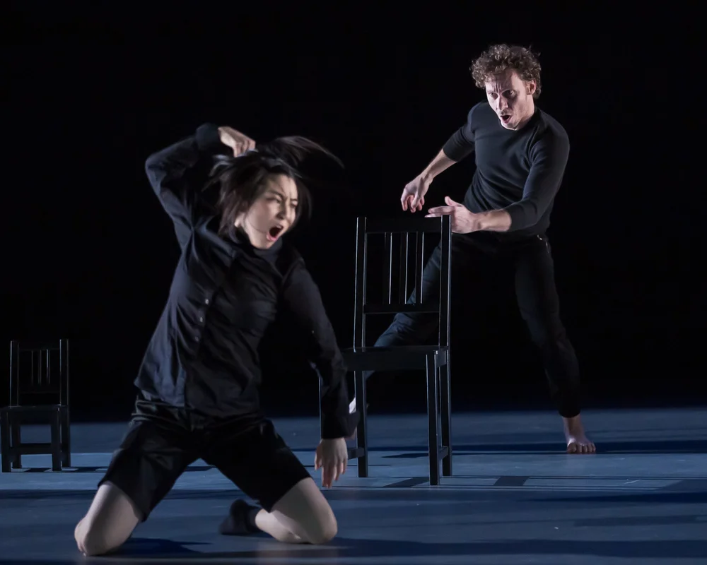  Sahara Morimoto and Ric Brown mid-movement. Sahara is kneeling with her legs spread shoulder-width apart and her right hand raised above her head. She appears to be yelling. Ric is behind her, and behind a chair stepping to the right and leaning rig