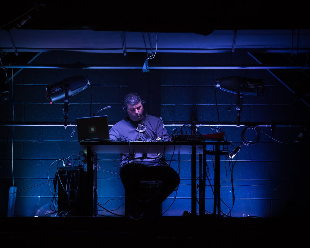  John Kameel Farah seated at a table, surrounded by sound equipment, seated in front of a blue black cinder block wall. 