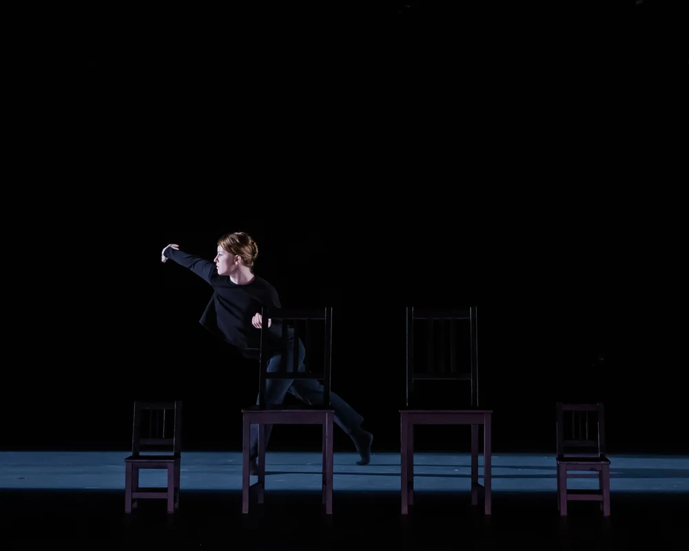  Sarah Fregeau mid-step, behind a line of four chairs. Her left foot is bent and crossed in front of her right, which is extended straight behind her.&nbsp; 