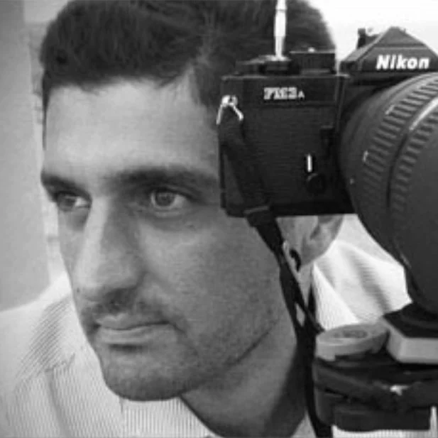 A photo of a man in black and white. He is using a camera and has short hair and a beard. He ears a white shirt.