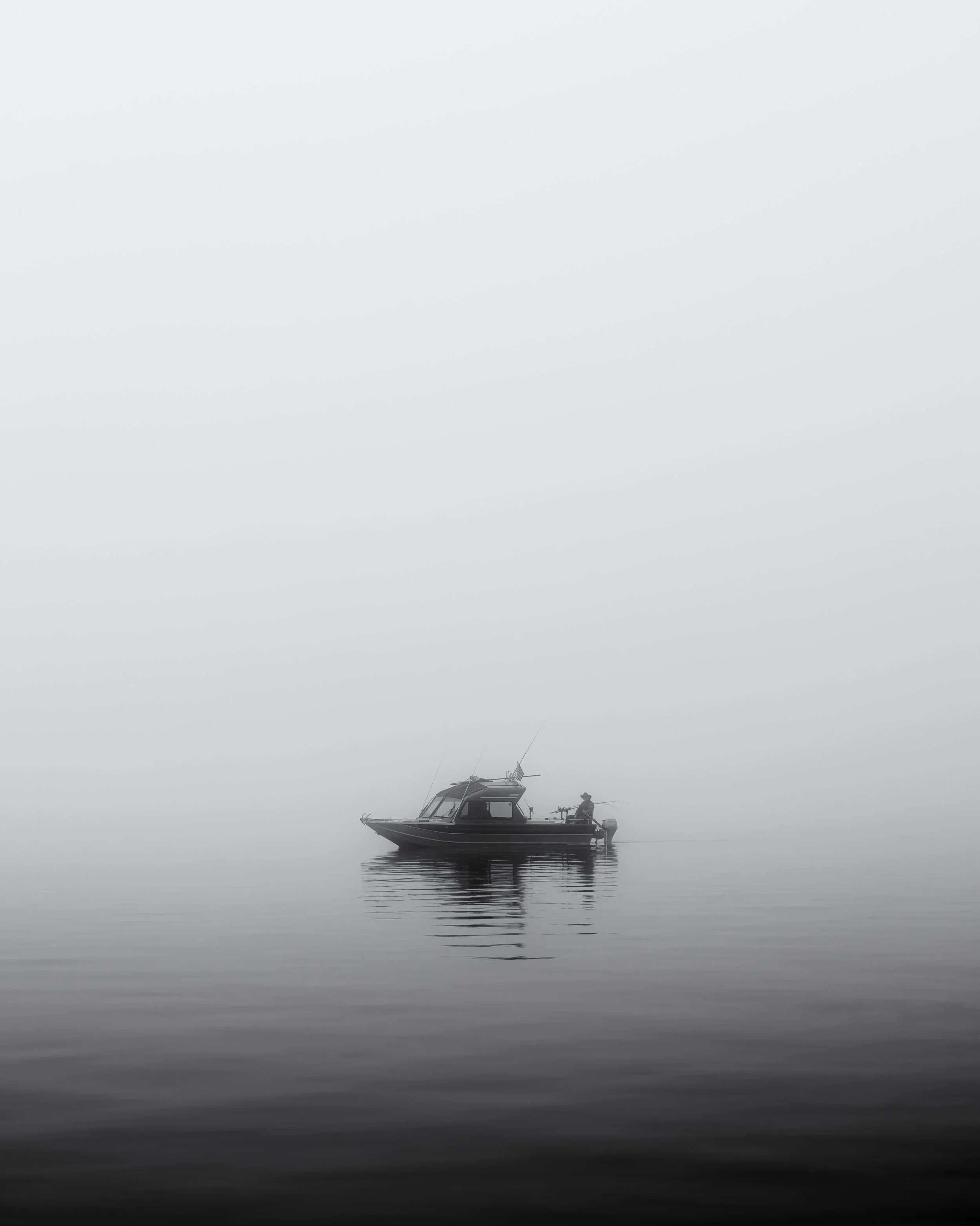 A small boat on calm water with a fisherman sitting at the edge, surrounded by fog.