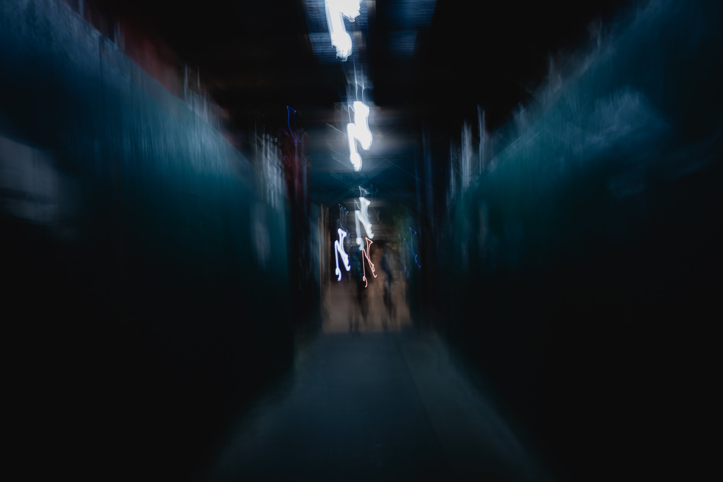 Blurred image of a dark alleyway with neon lights and two people walking in the distance.