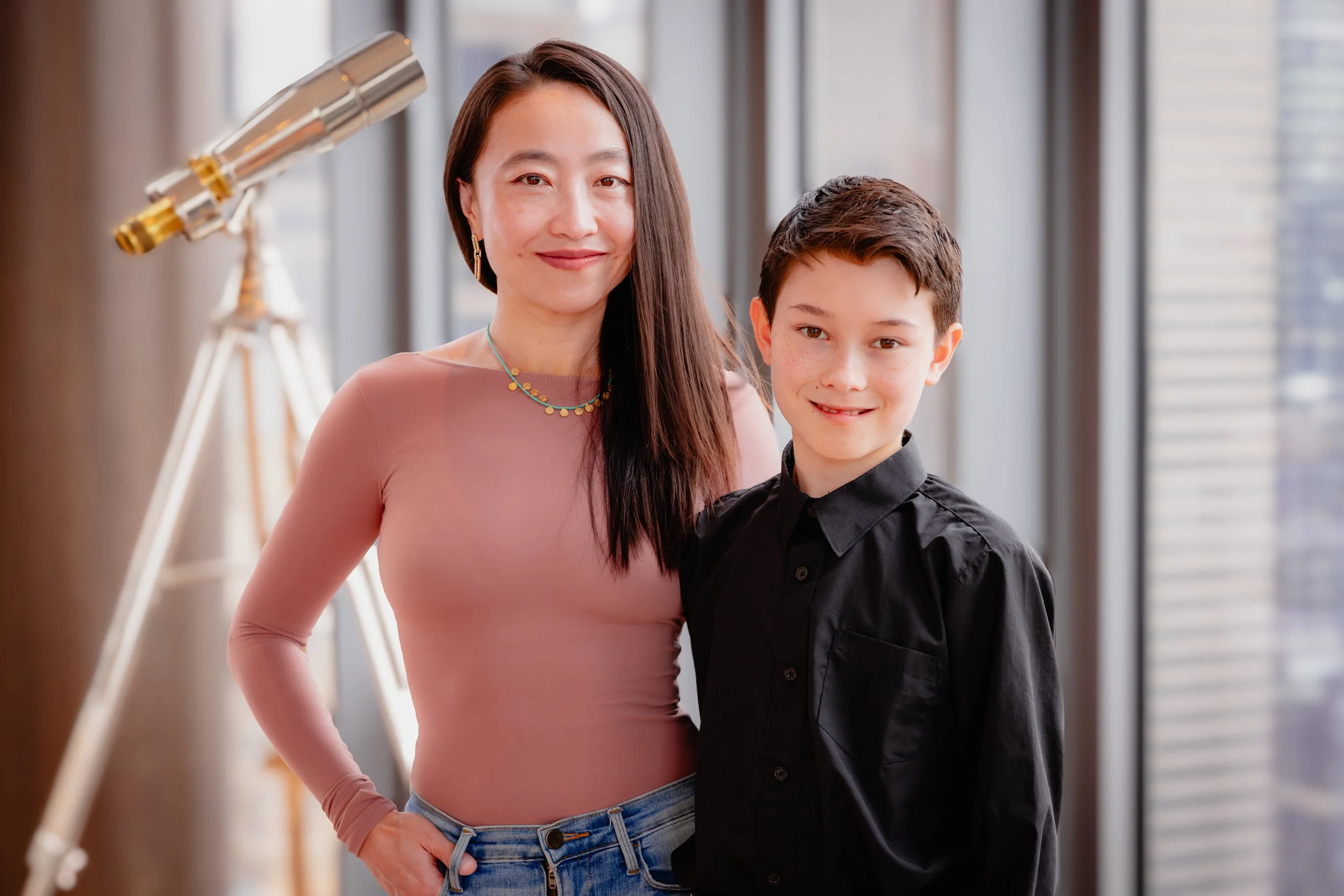 A smiling woman and boy standing indoors near large windows, with a telescope in the background.