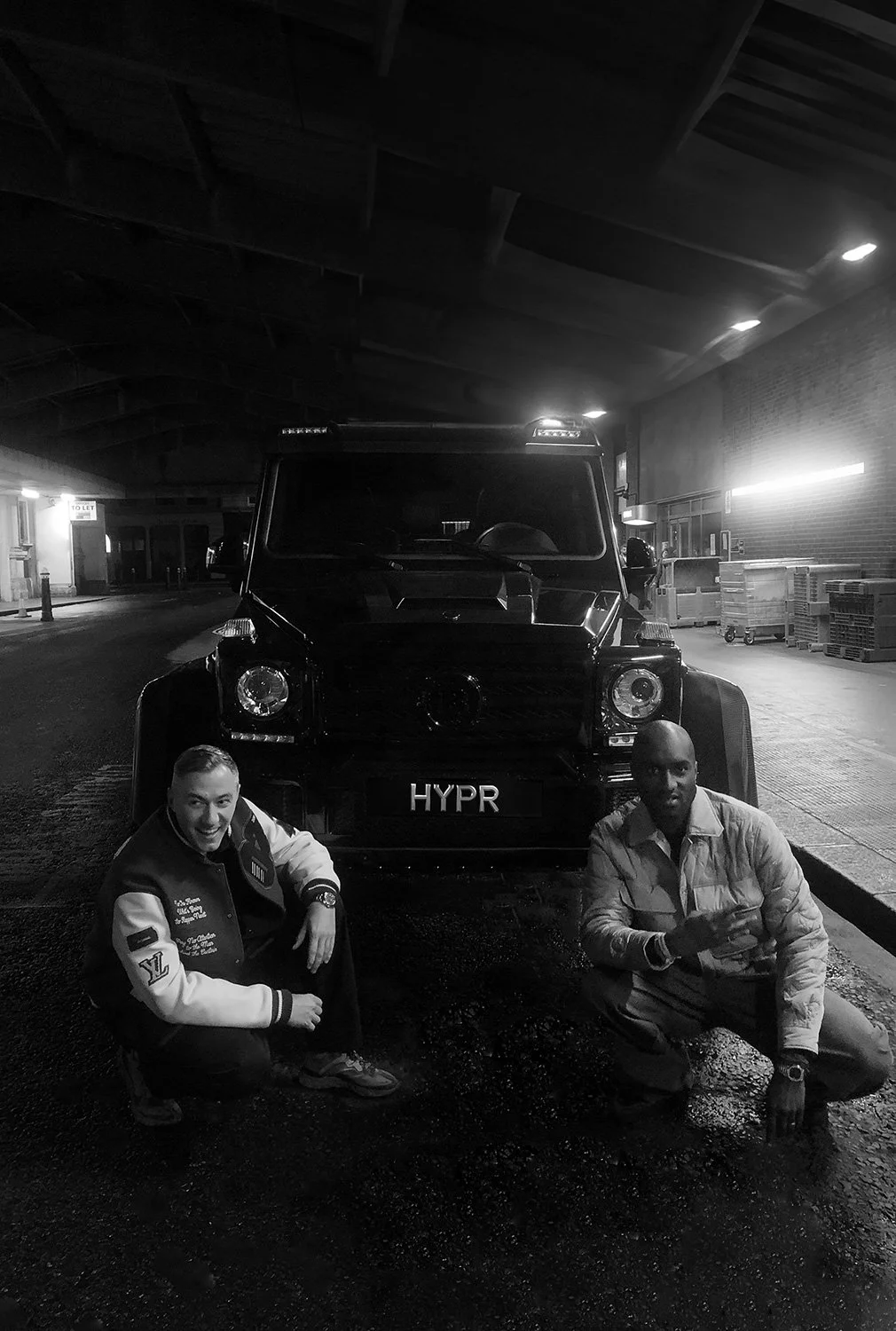 Two men squatting in front of a black vehicle with a license plate that reads 'HYPR' inside a dimly lit area under a roof, with some stacked pallets and a building in the background.