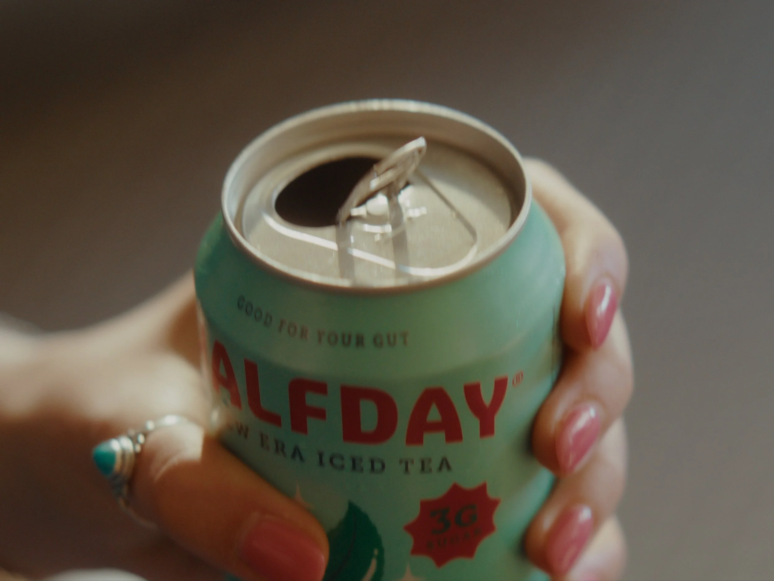 Close-up of a hand holding an aluminum can of iced tea with the can tab pulled back, revealing the opening.
