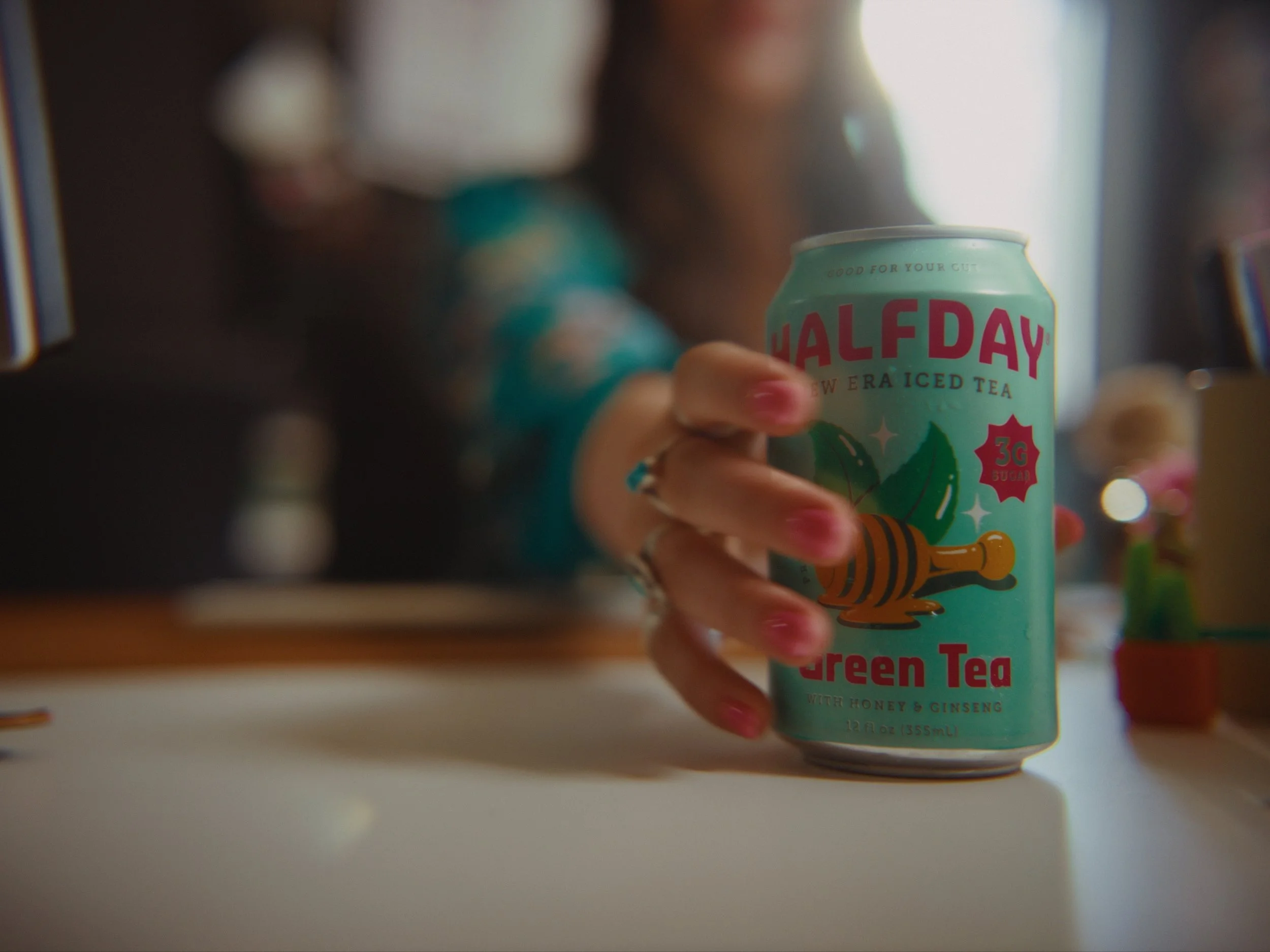 A hand holding a can of Halcy Green Tea with honey and ginseng, with a blurred background of other cans on a store shelf.