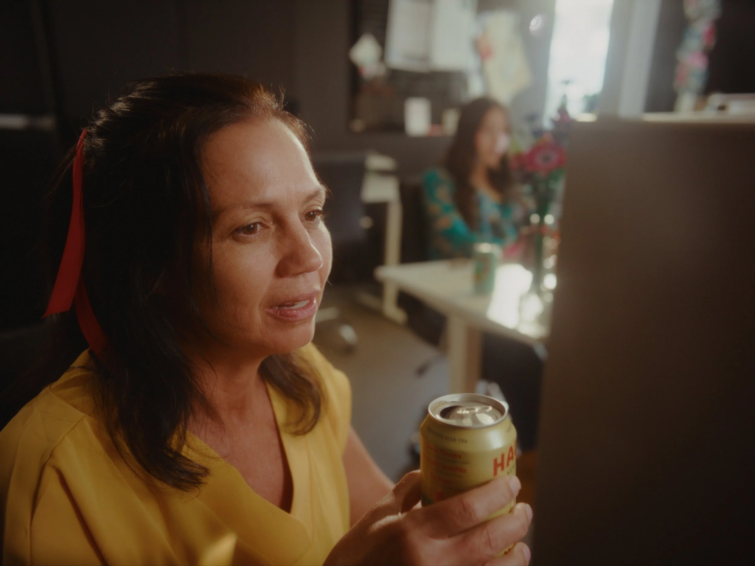 A woman in a yellow top holding a can of Campbell's Chunky soup, sitting in a room with a blurred background including another woman in a blue top and various objects.