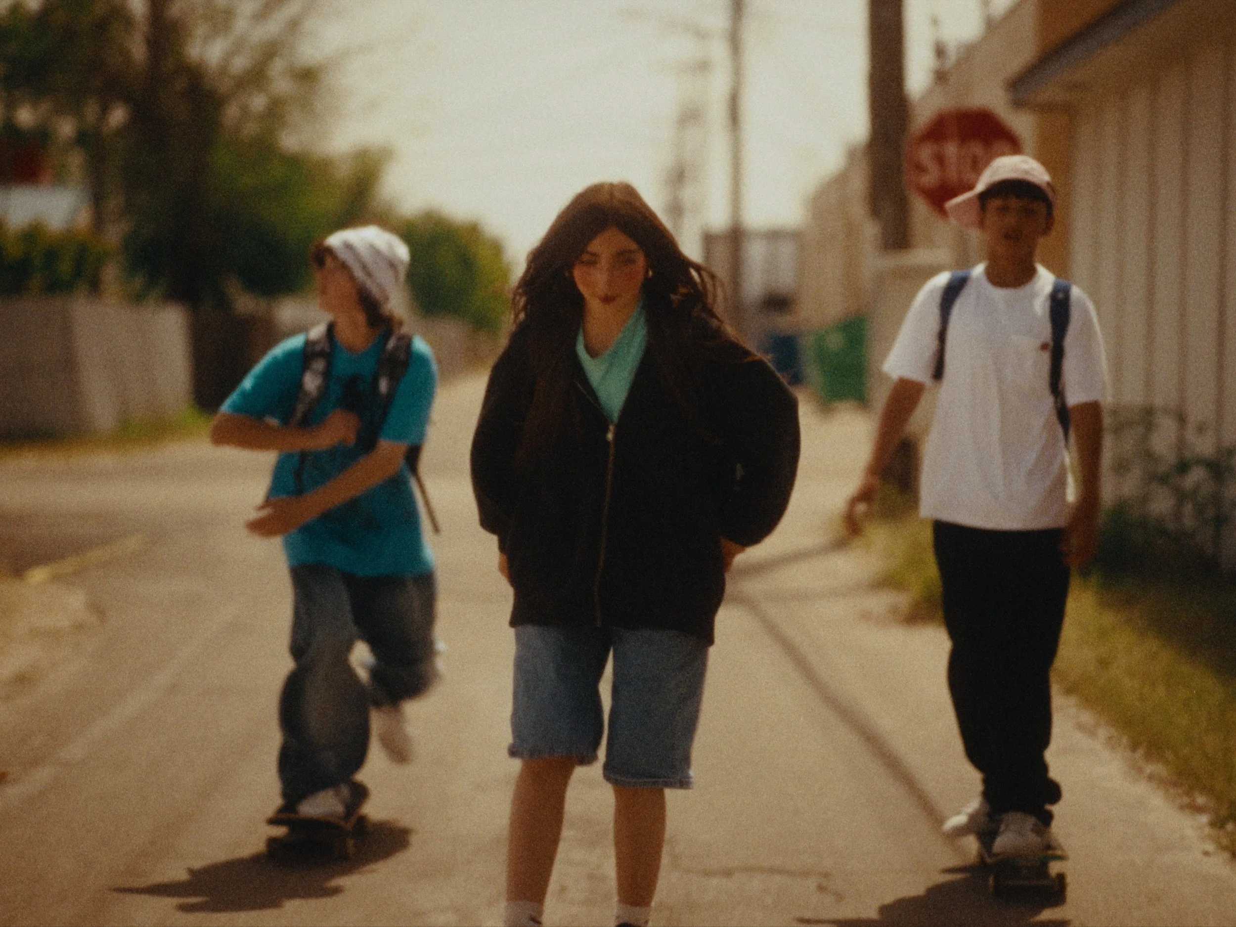 Three children walking outdoors on a sunny day, two boys and a girl, wearing casual clothes and backpacks.