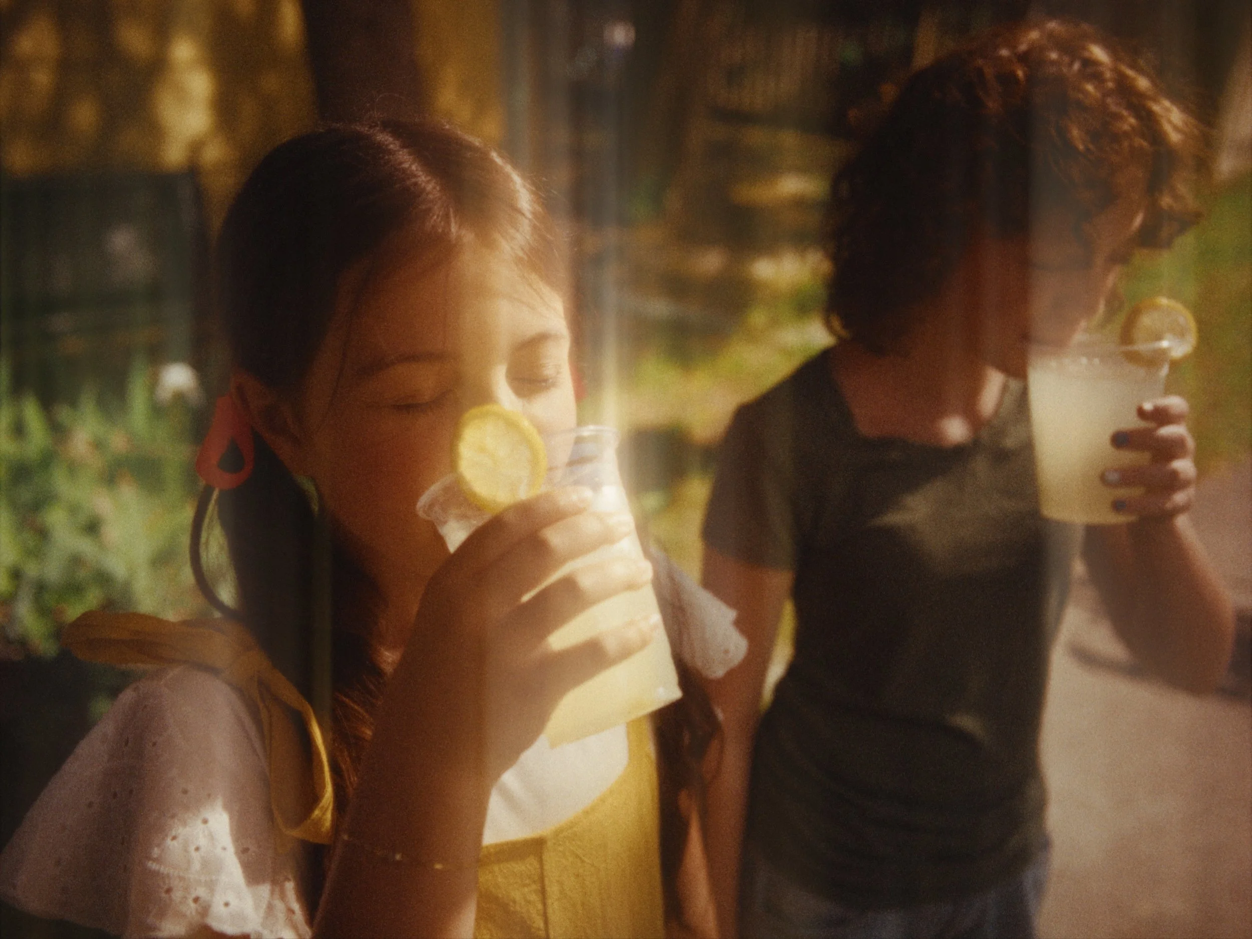 Two young girls sitting outdoors, enjoying drinks with lemon slices, during daytime.