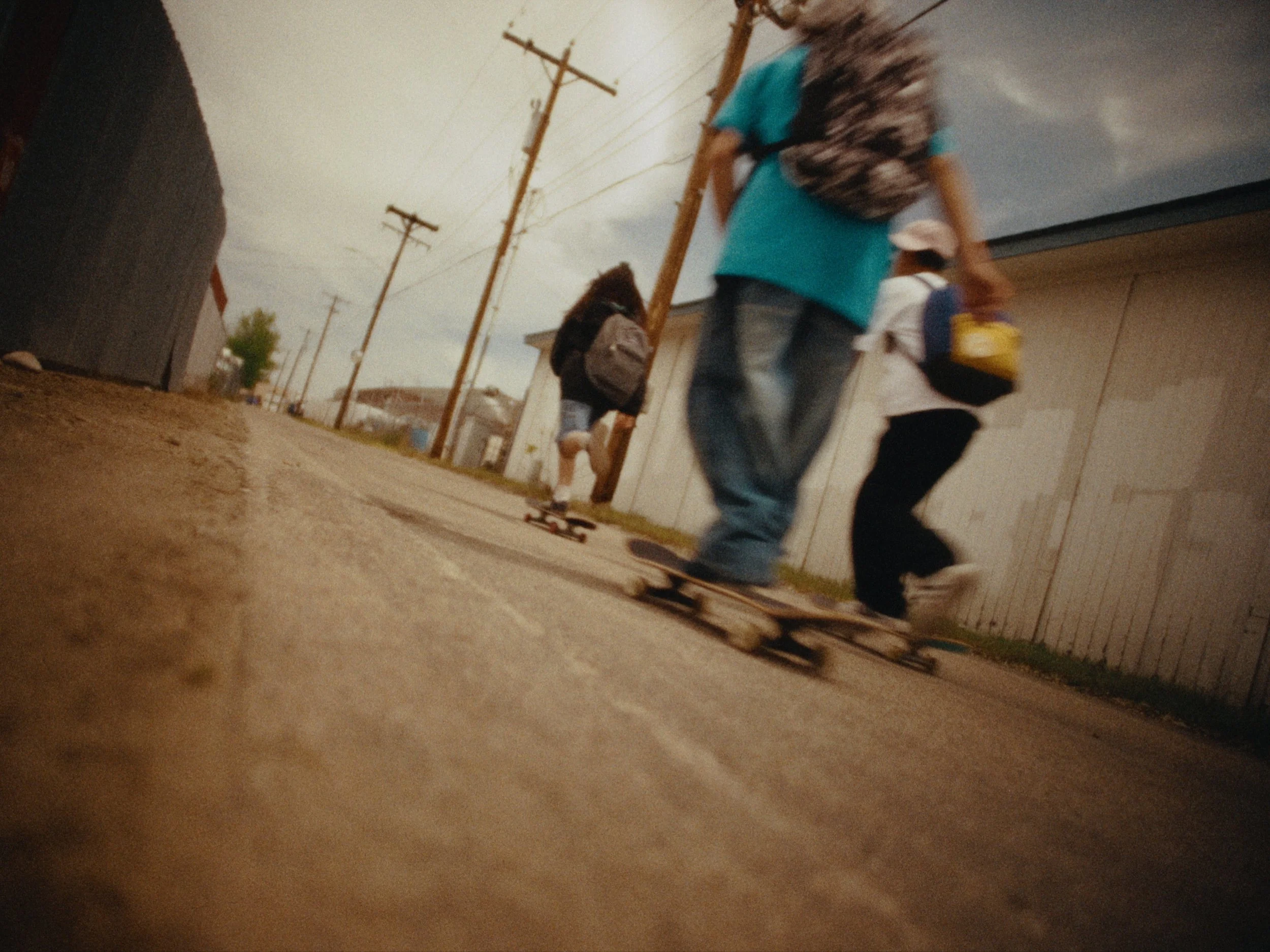 A person riding a skateboard on a dirt path, wearing a blue shirt and carrying a backpack. Several other people are walking or skateboarding along the same path. Wooden utility poles and a building are visible in the background under an overcast sky.
