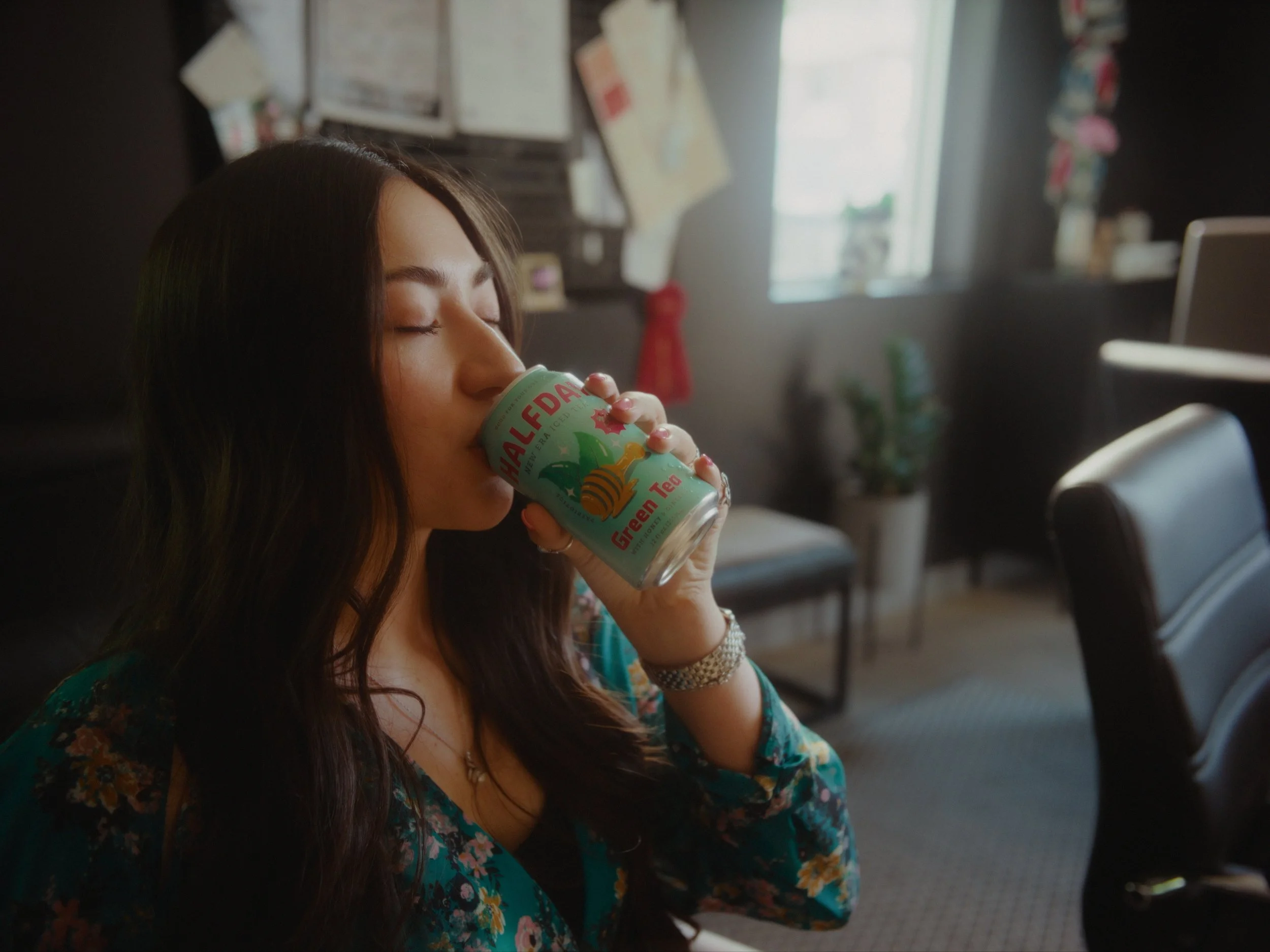 Young woman with long dark hair drinking from a can of Hawaiian Green Tea, sitting indoors with blurred background of a window and office or home decor.