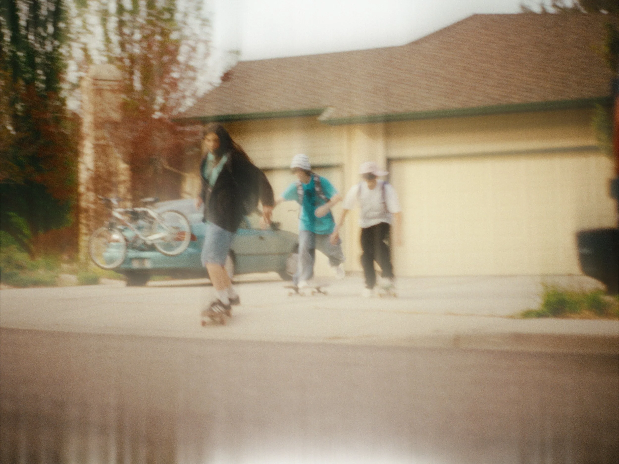Three young women walking outside near a garage, one carrying a bike, in a suburban neighborhood.