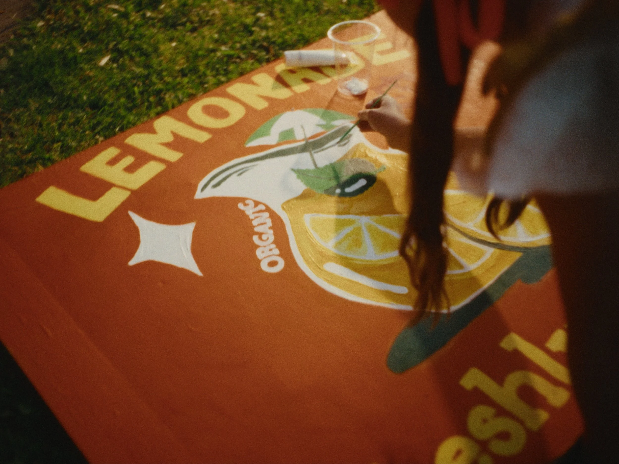 A person painting a colorful sign on a board with the words 'WELCOME' and images of lemon slices and a lime, outdoors on grass.