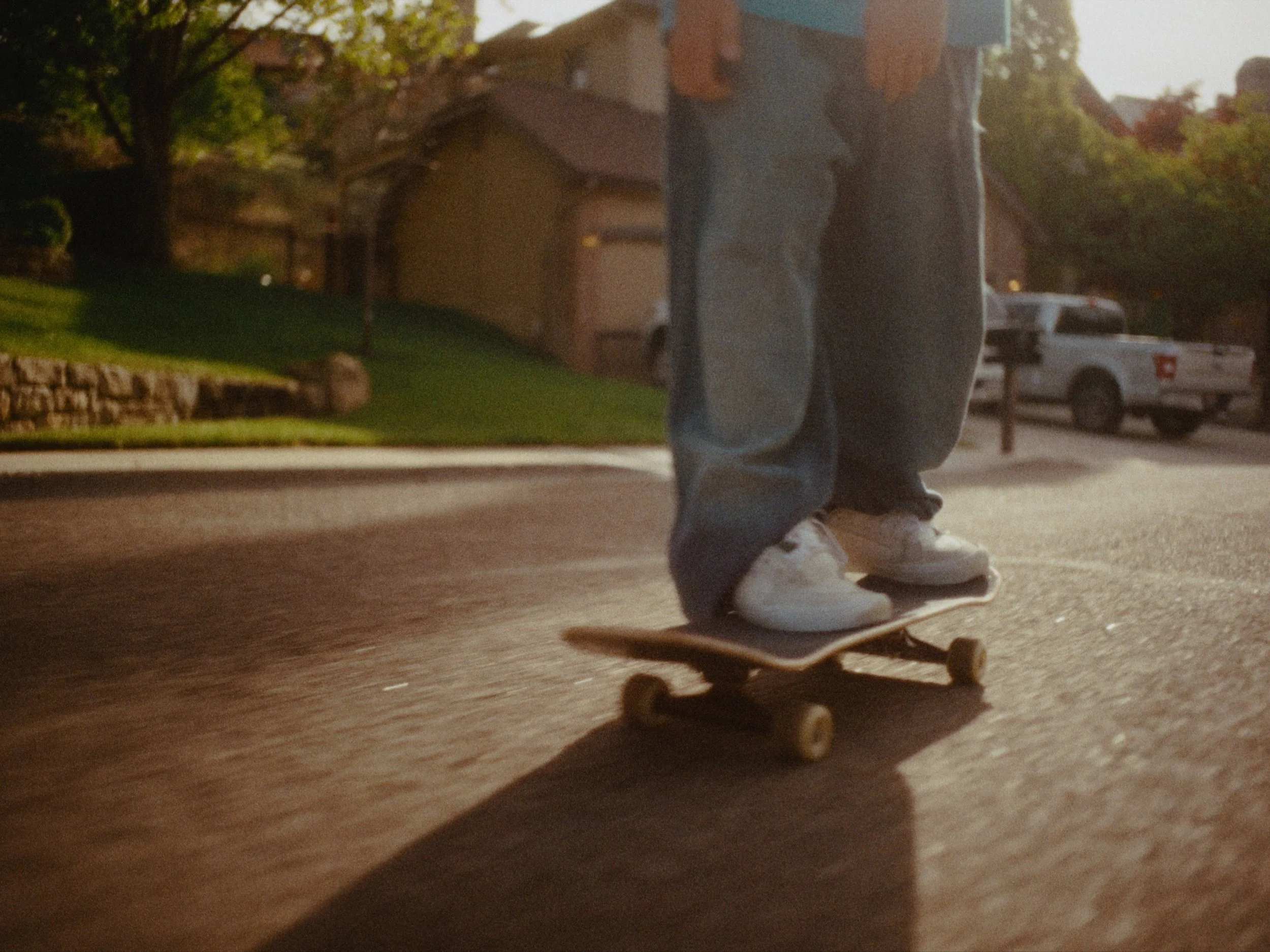 A person skateboarding on a sidewalk in a suburban neighborhood during daylight. The person is wearing jeans and white sneakers, with trees, houses, and a parked pickup truck visible in the background.